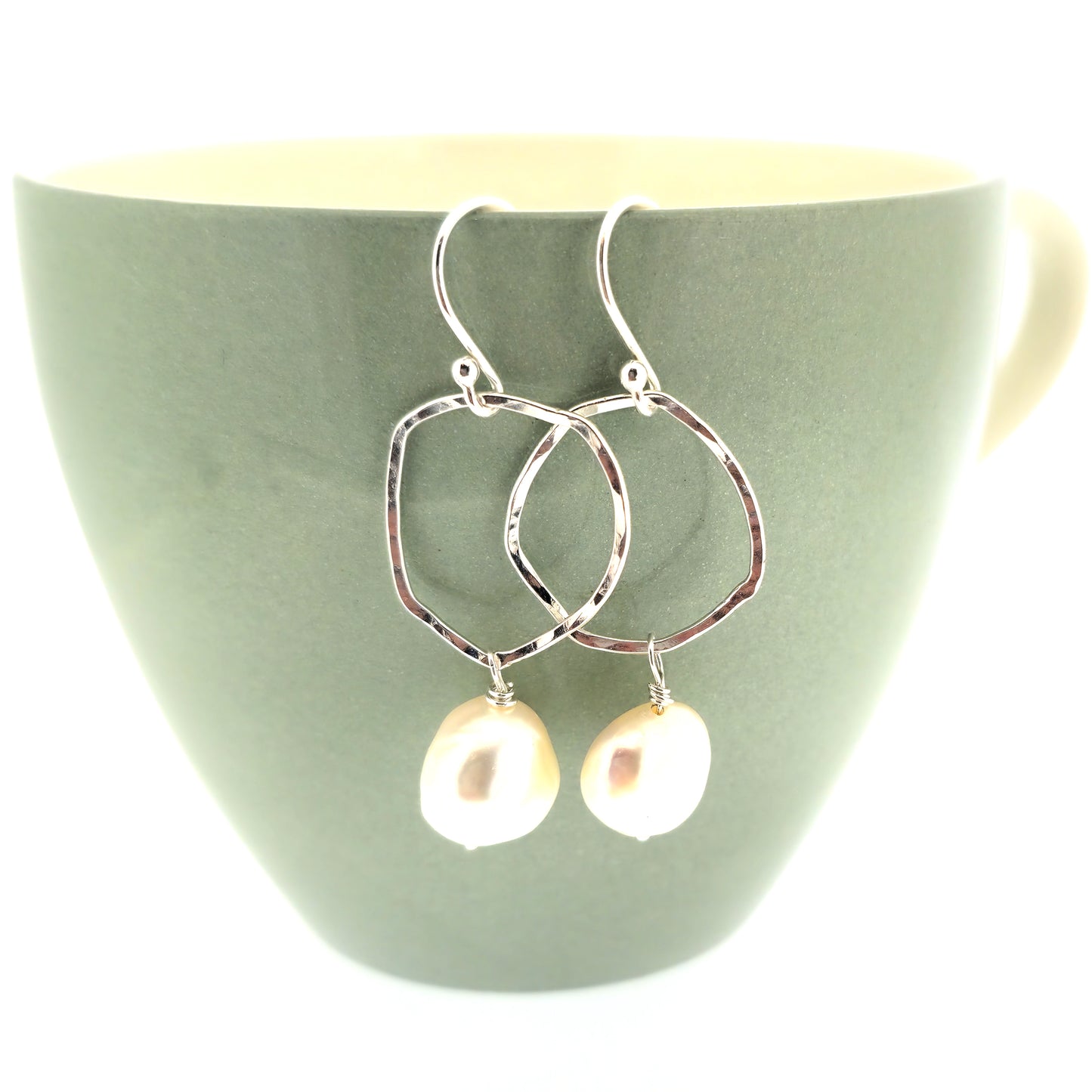 Silver drop earrings consisting of an organic rustic style open circle with a hammered finish suspended from silver ear hooks and with a white pearl hanging from it. Large. Pictured on a cup.