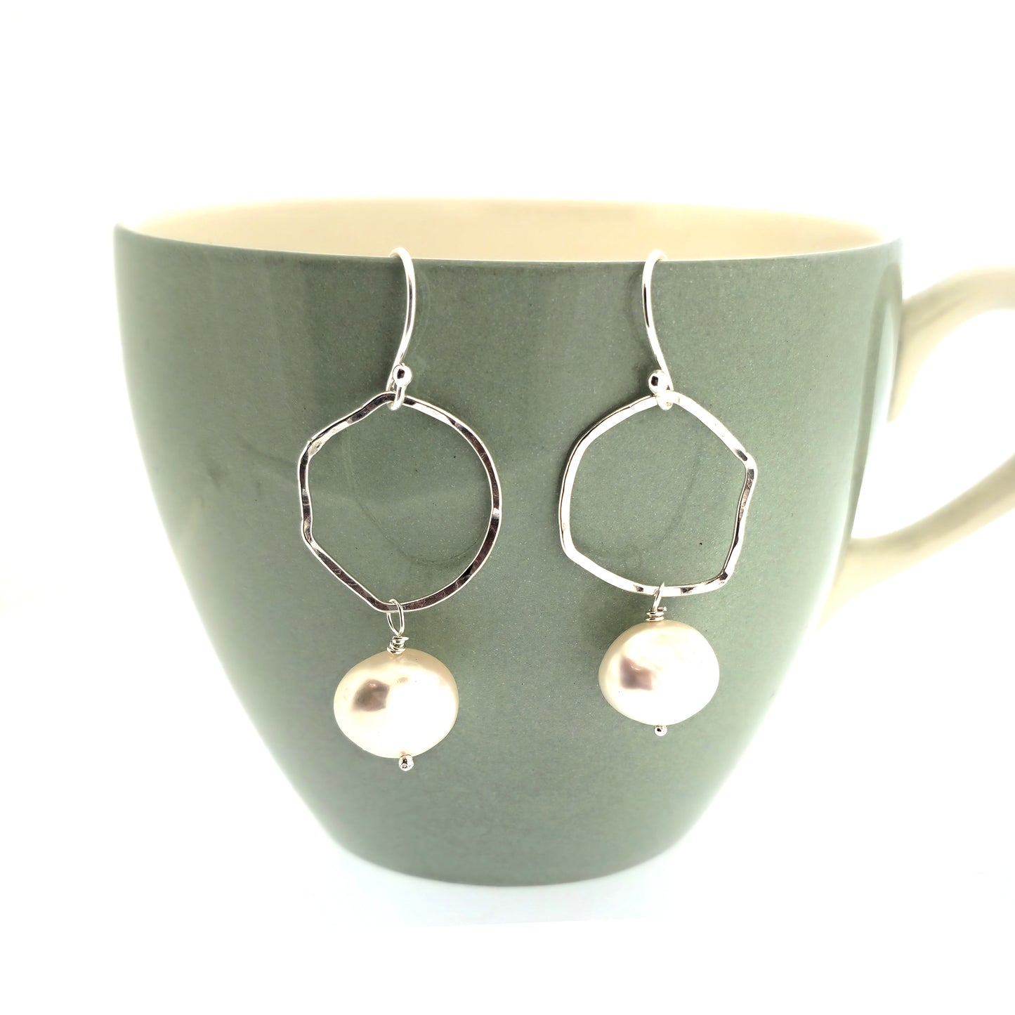 Silver drop earrings consisting of an organic rustic style open circle with a hammered finish suspended from silver ear hooks and with a white pearl hanging from it. Large. Pictured on a cup.