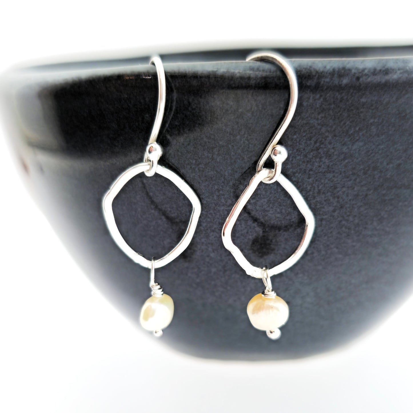 Silver drop earrings consisting of an organic rustic style open circle with a hammered finish suspended from silver ear hooks and with a white pearl hanging from it. Small. Pictured on a black bowl.