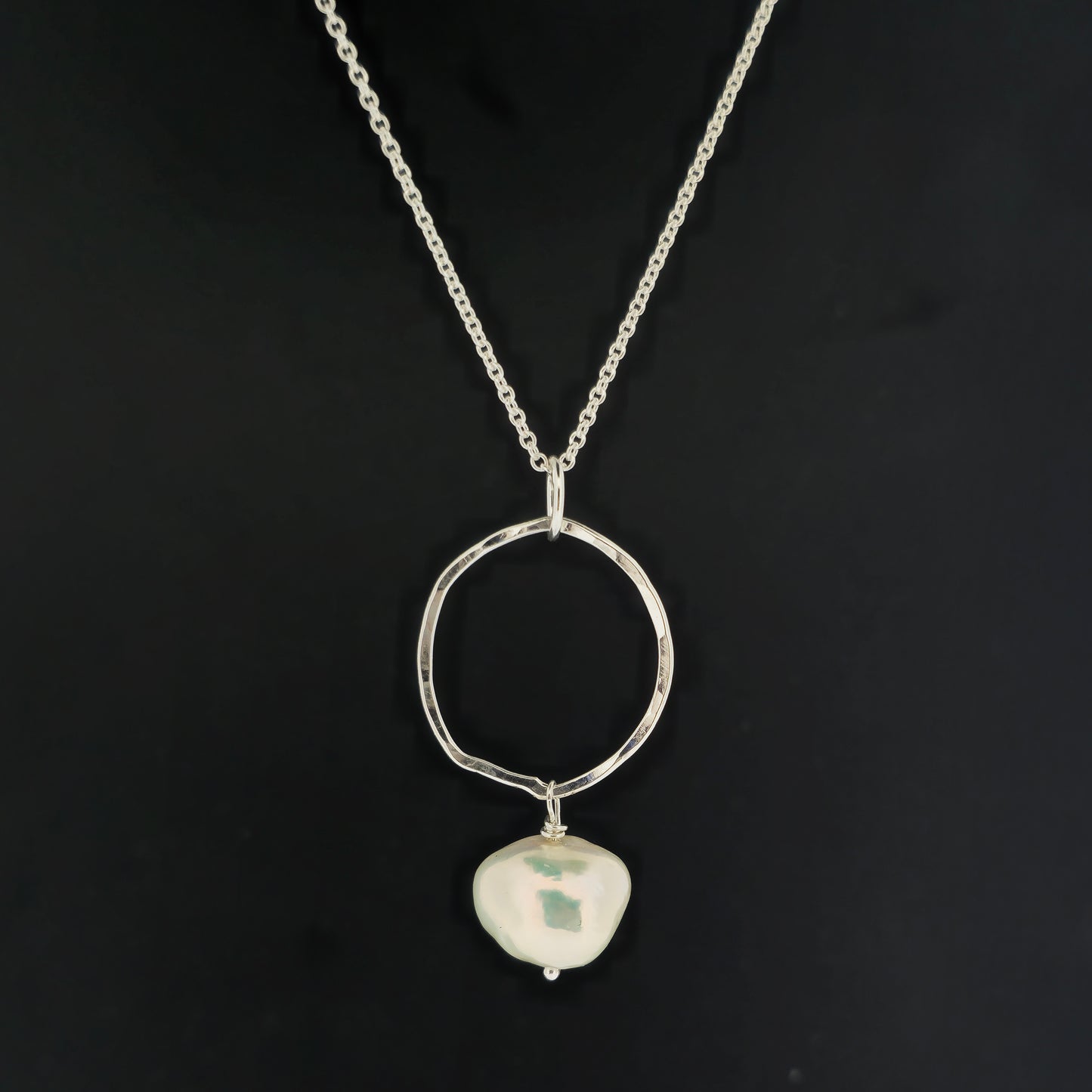 A silver pendant consisting of an organic rustic style open circle with a hammered finish and a pearl suspended from it. Hanging from a silver chain. Large. Pictured on a black background.