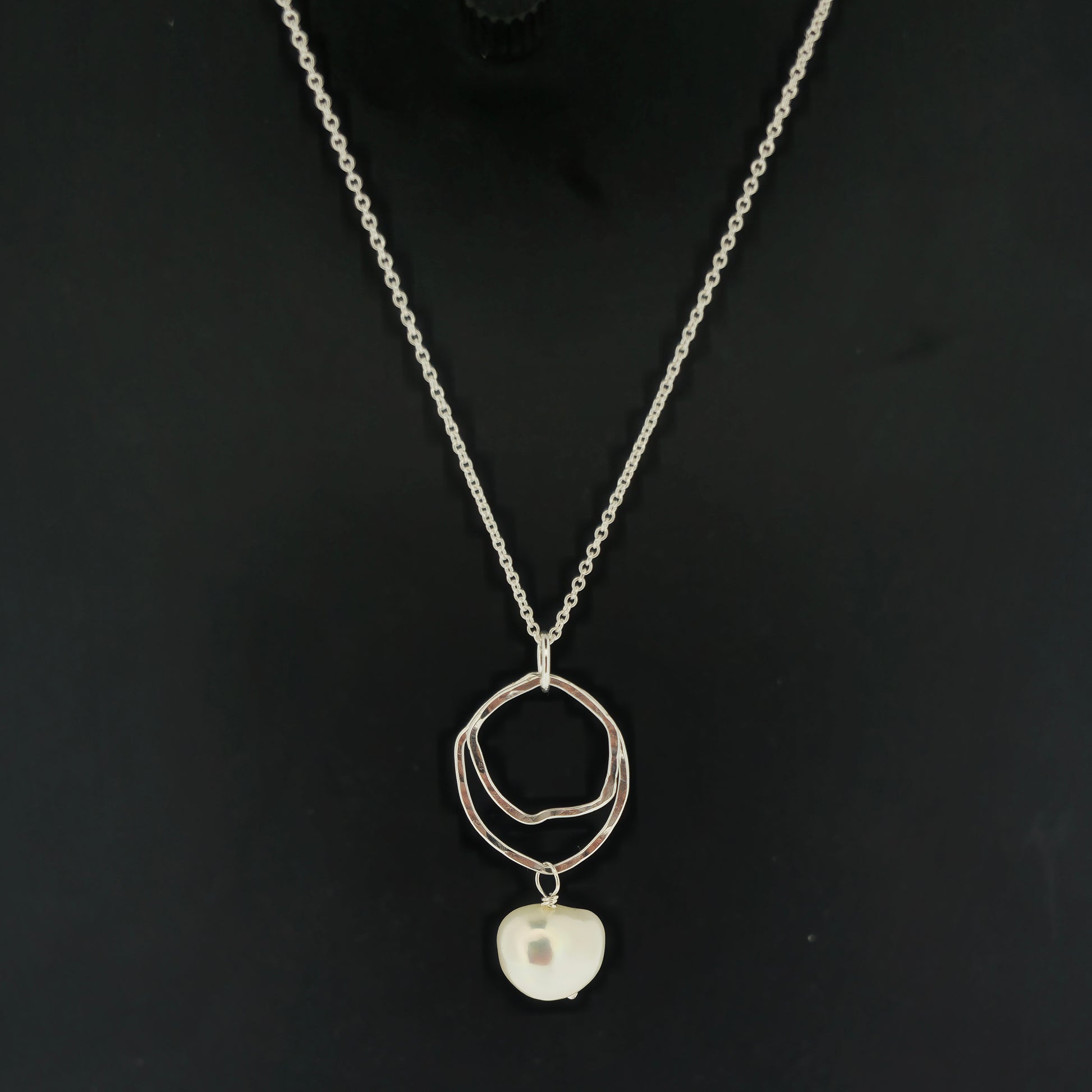 A silver pendant consisting of two organic rustic style open circles of different sizes with a hammered finish and an ivory pearl suspended beneath. Hanging from a silver chain. On a black background.