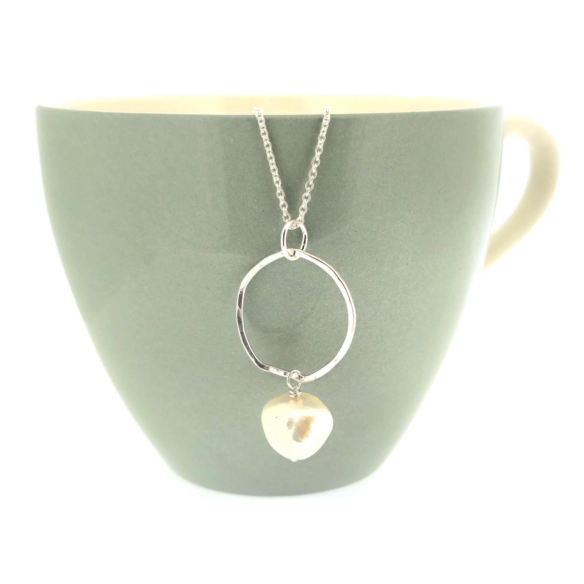 A silver pendant consisting of an organic rustic style open circle with a hammered finish and a pearl suspended from it. Hanging from a silver chain. Large. Pictured on a mug.