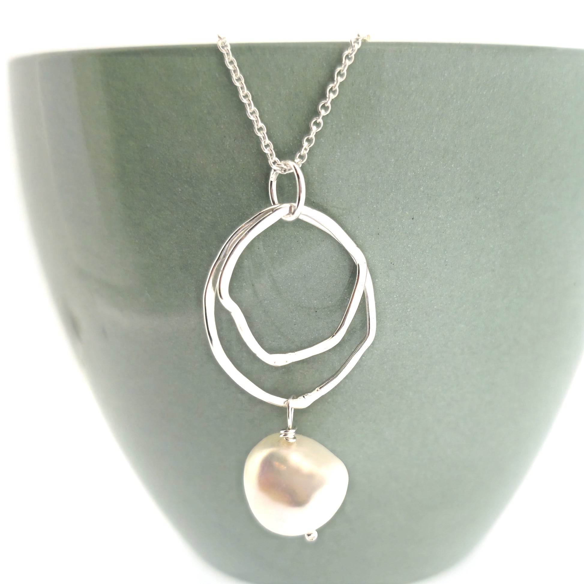 A silver pendant consisting of two organic rustic style open circles of different sizes with a hammered finish and an ivory pearl suspended beneath. Hanging from a silver chain. Pictured on a cup.