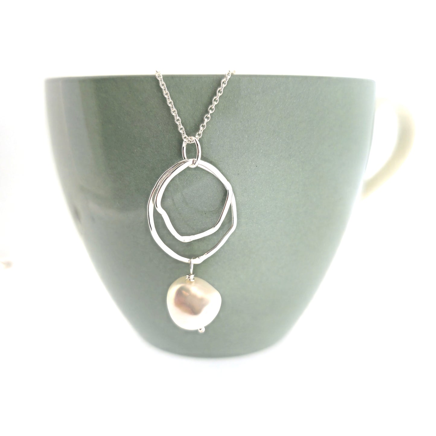 A silver pendant consisting of two organic rustic style open circles of different sizes with a hammered finish and an ivory pearl suspended beneath. Hanging from a silver chain. Pictured on a cup.