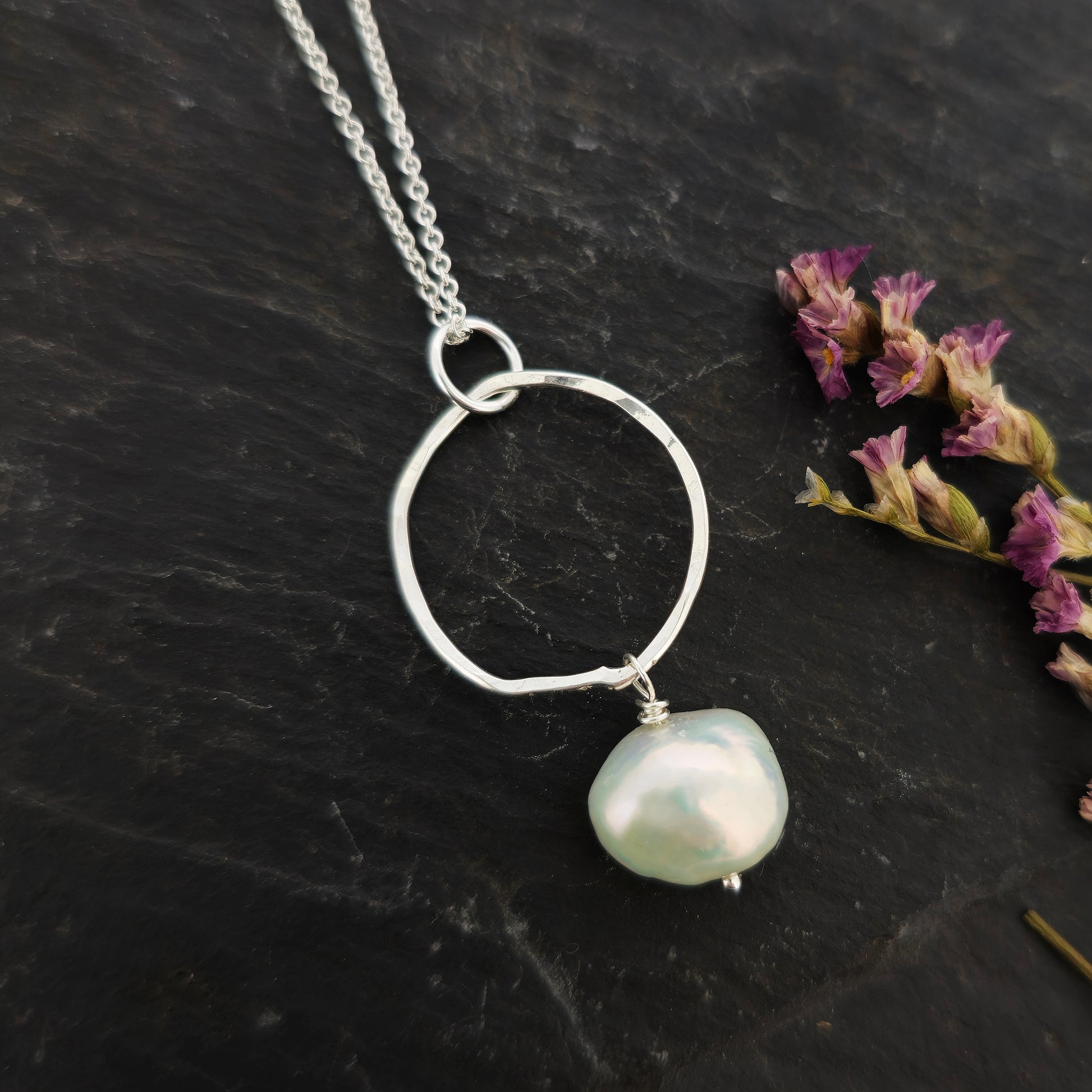 A silver pendant consisting of an organic rustic style open circle with a hammered finish and a pearl suspended from it. Hanging from a silver chain. Large. Pictured on slate with flowers.