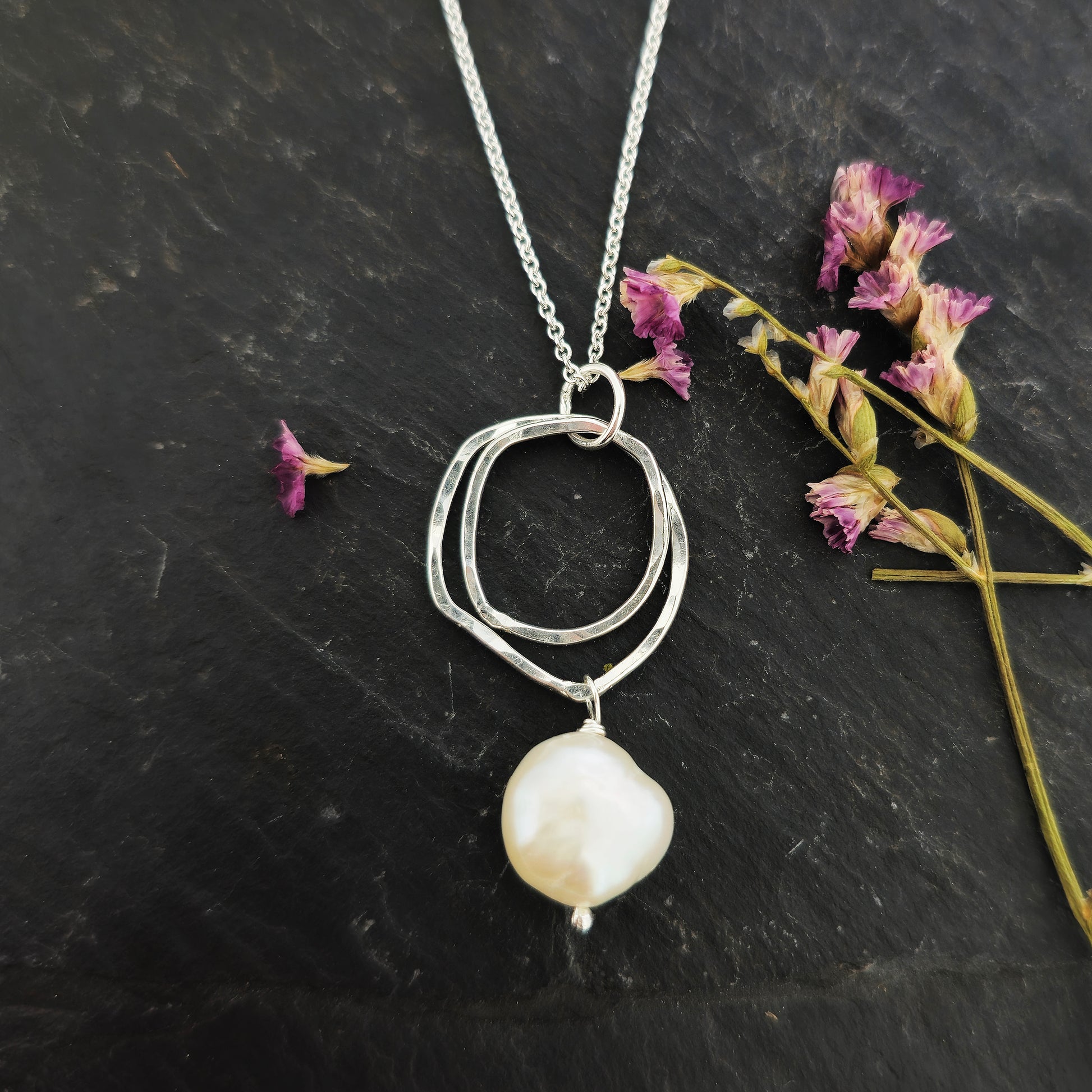 A silver pendant consisting of two organic rustic style open circles of different sizes with a hammered finish and an ivory pearl suspended beneath. Hanging from a silver chain. Pictured on slate with flowers.