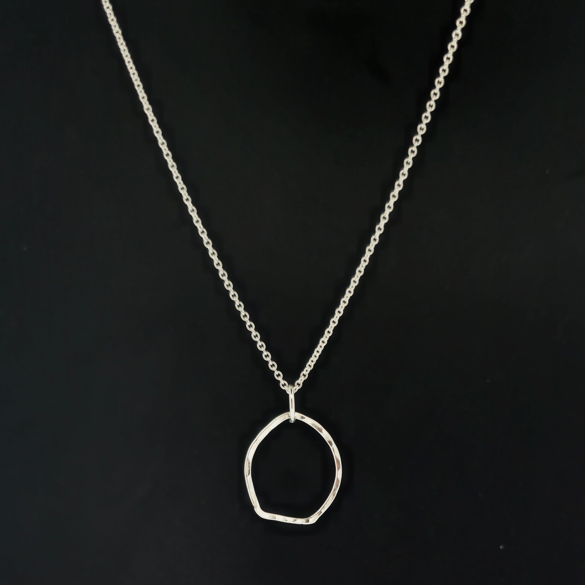 A silver pendant necklace consisting of an open organic rustic style silver circle with a hammered finish suspended from a silver chain - large. On a black background.