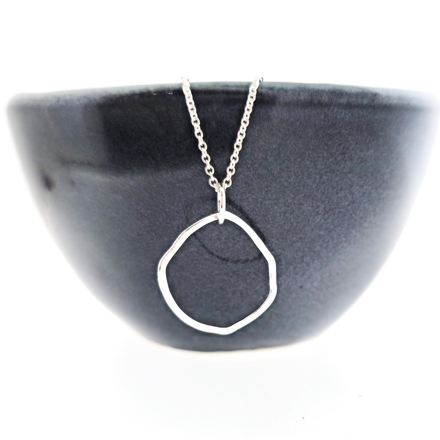 A silver pendant necklace consisting of an open organic rustic style silver circle with a hammered finish suspended from a silver chain - large. Pictured on a black bowl.