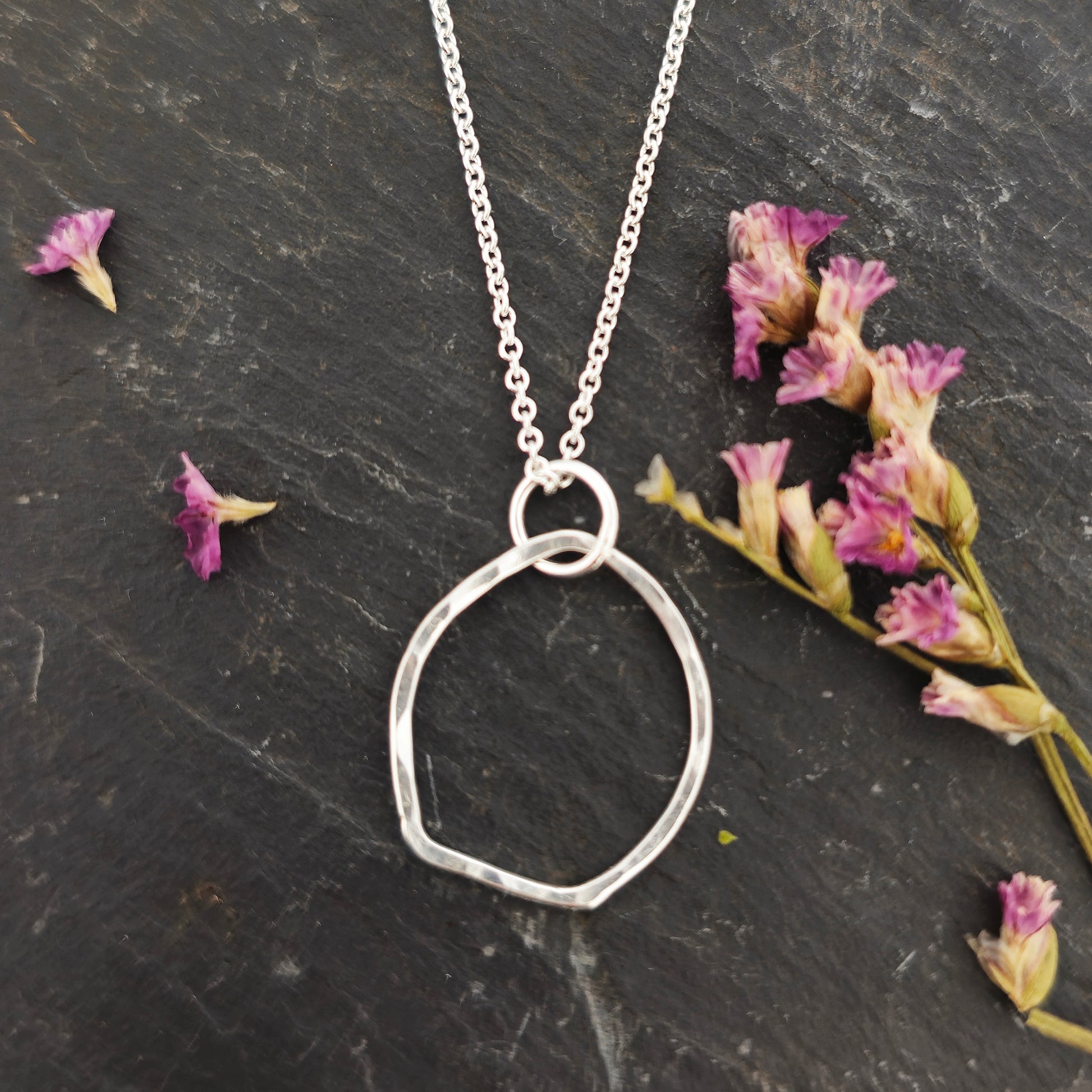 A silver pendant necklace consisting of an open organic rustic style silver circle with a hammered finish suspended from a silver chain - large. Pictured on slate with flowers.