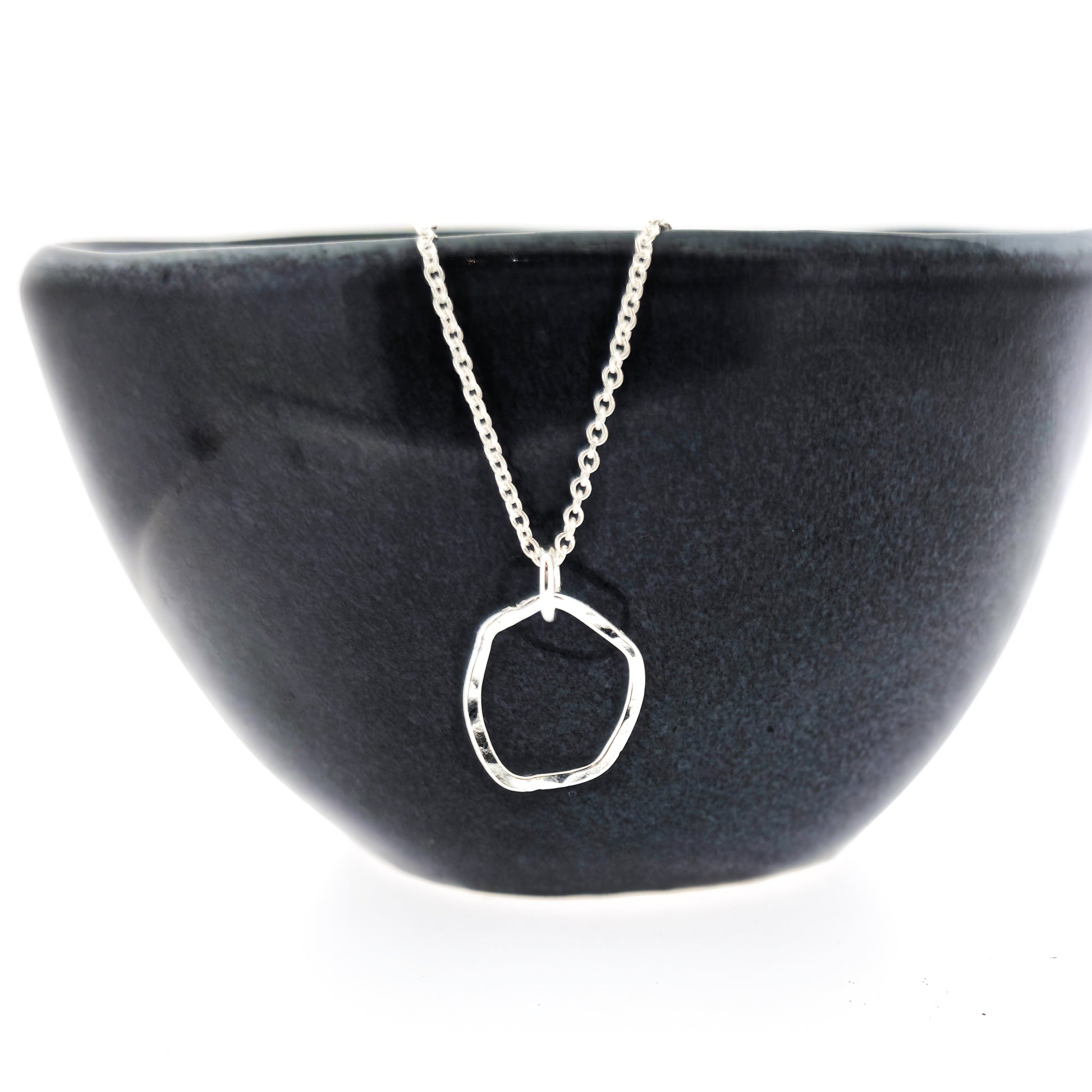 A silver pendant necklace consisting of an open organic rustic style silver circle with a hammered finish suspended from a silver chain - small. Pictured on a black bowl.