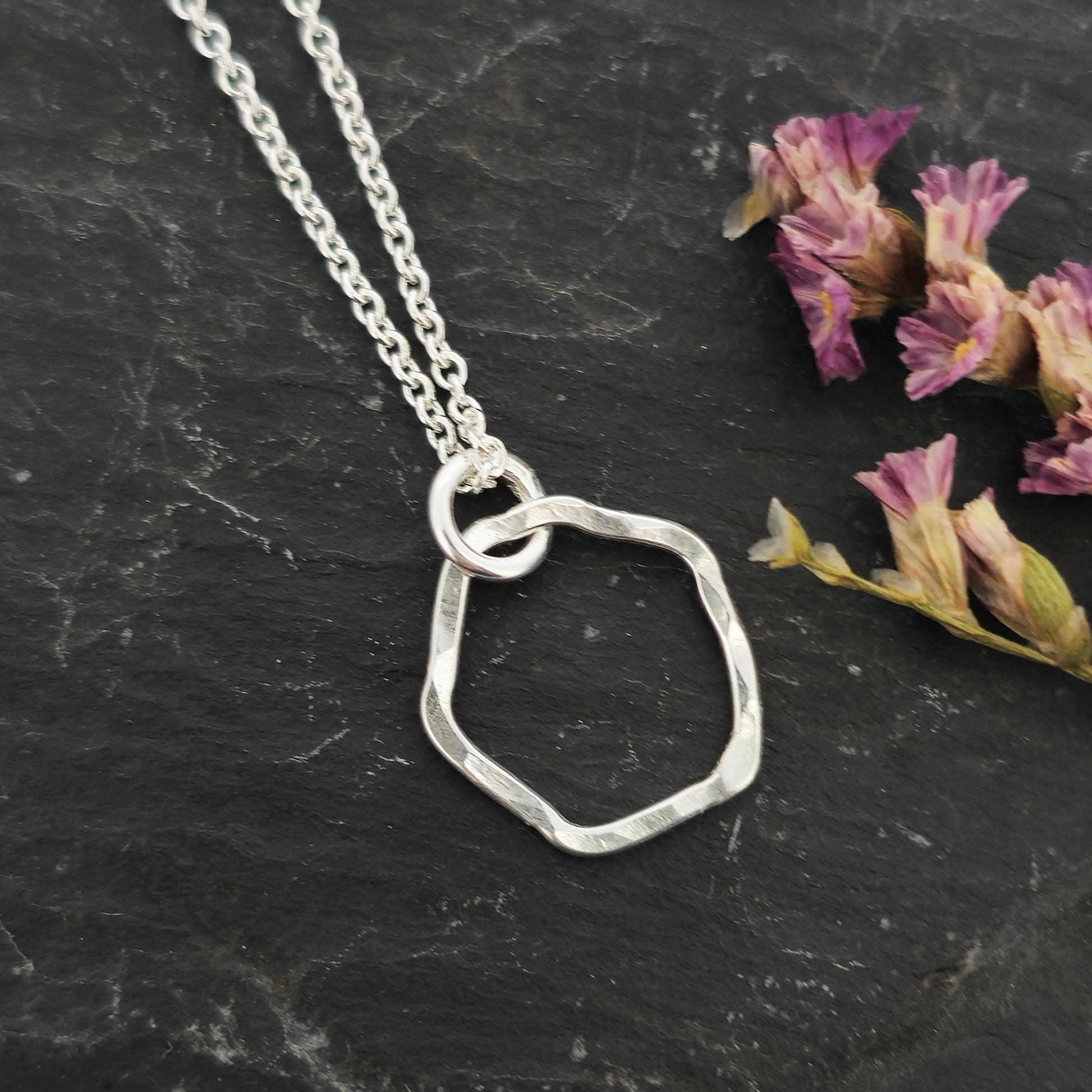 A silver pendant necklace consisting of an open organic rustic style silver circle with a hammered finish suspended from a silver chain - small. Pictured on slate with flowers.