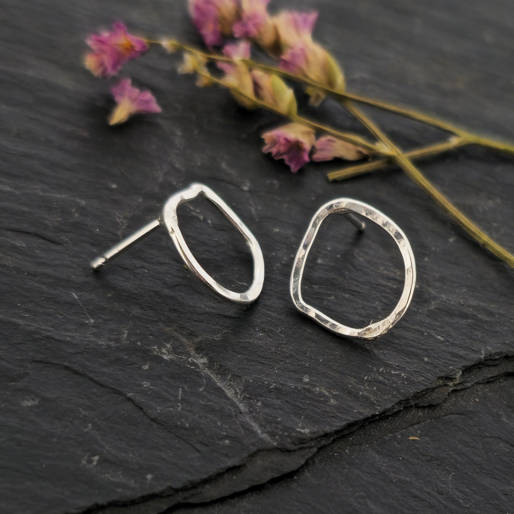 Silver open stud earrings of an organic rustic circle shape - small. Shown on slate with flowers.