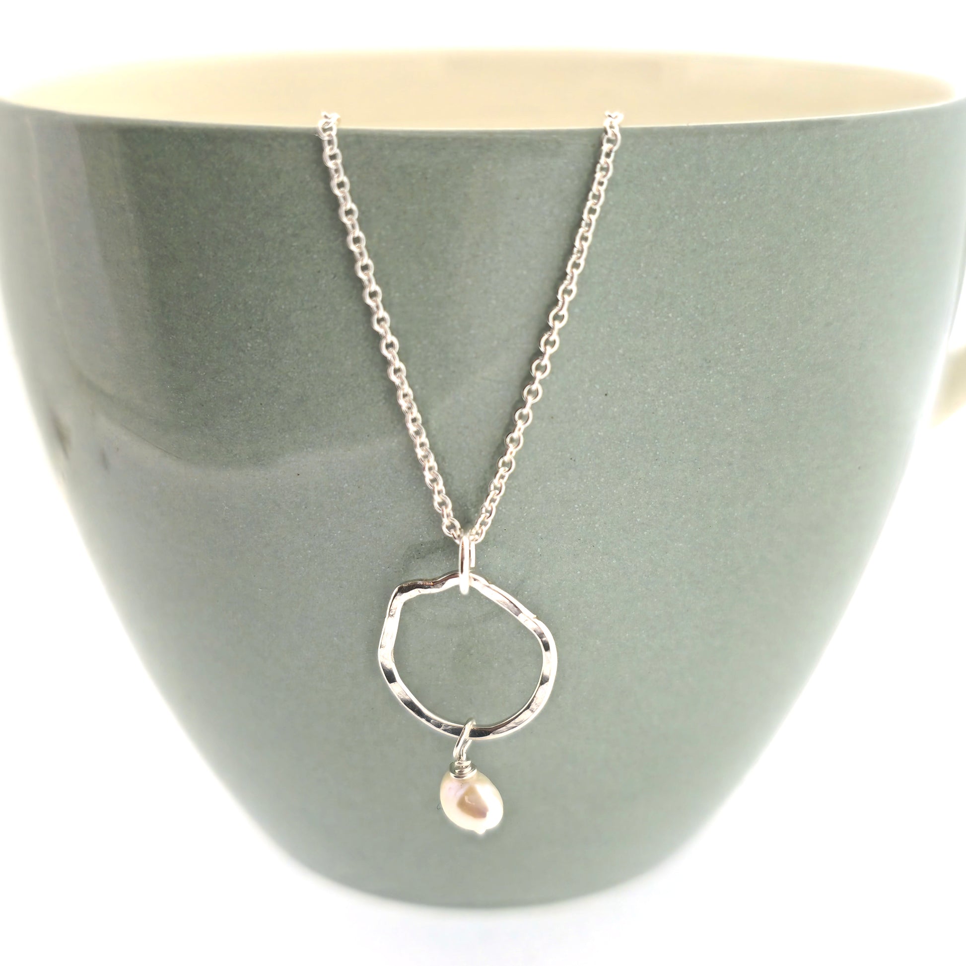 A silver pendant consisting of an organic rustic style open circle with a hammered finish and a pearl suspended from it. Hanging from a silver chain. Small. Pictured on a mug