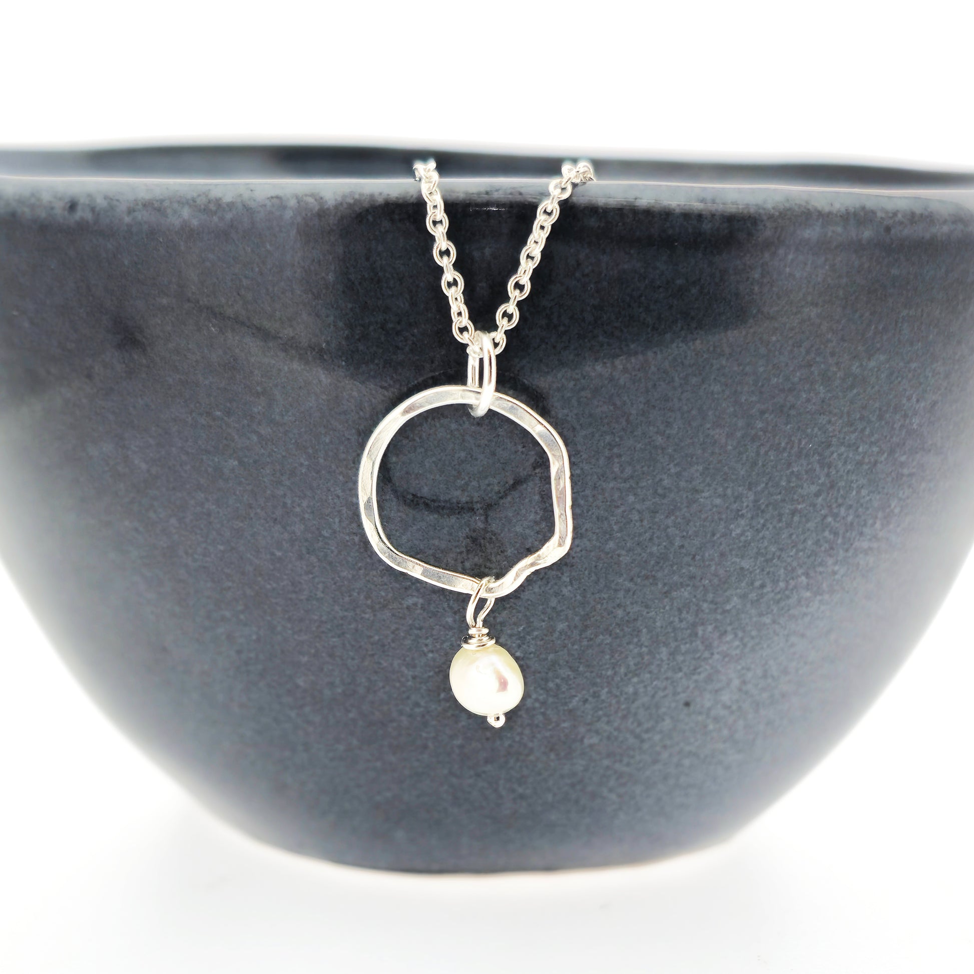 A silver pendant consisting of an organic rustic style open circle with a hammered finish and a pearl suspended from it. Hanging from a silver chain. Small. Pictured on a black bowl.