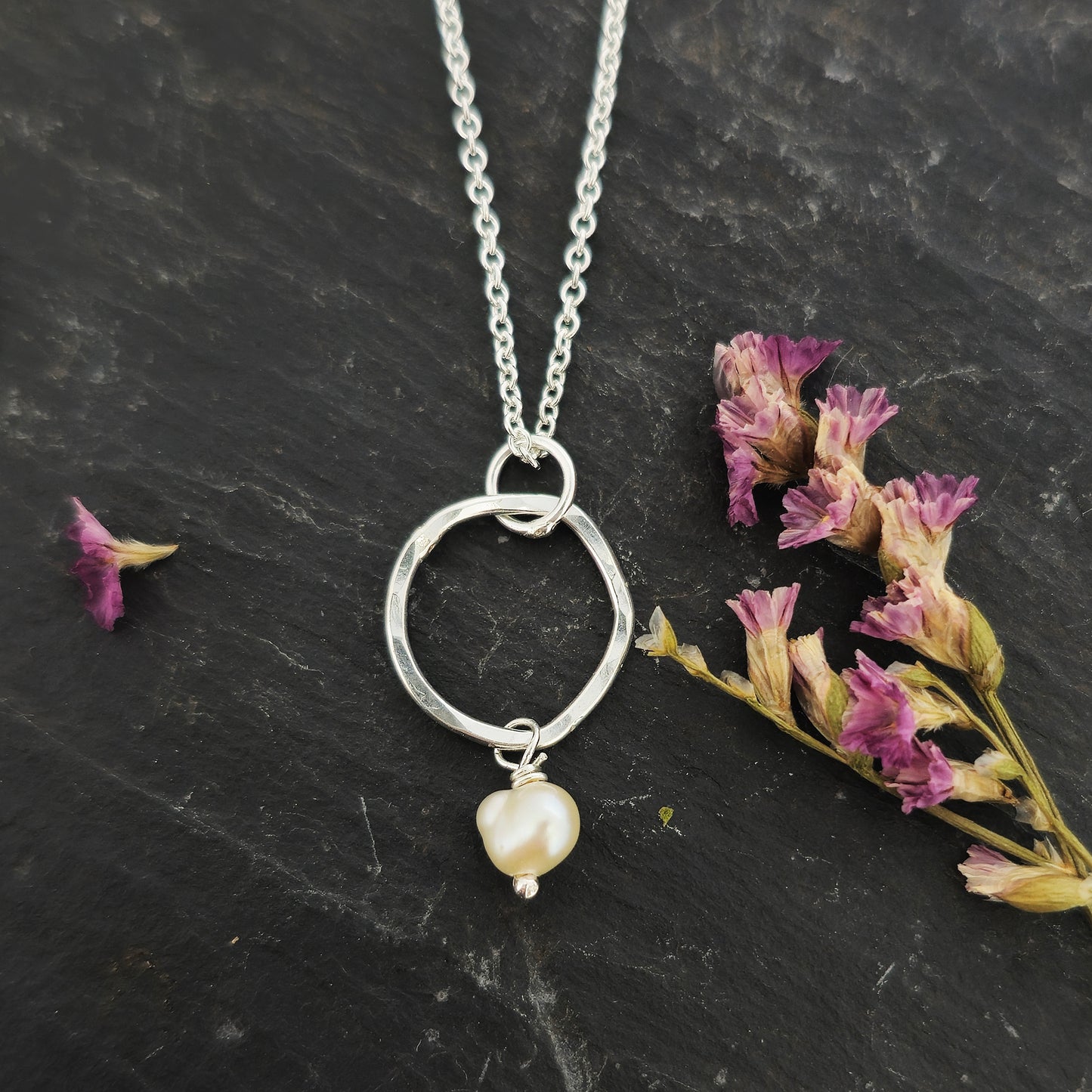 A silver pendant consisting of an organic rustic style open circle with a hammered finish and a pearl suspended from it. Hanging from a silver chain. Small. Pictured on slate with flowers.