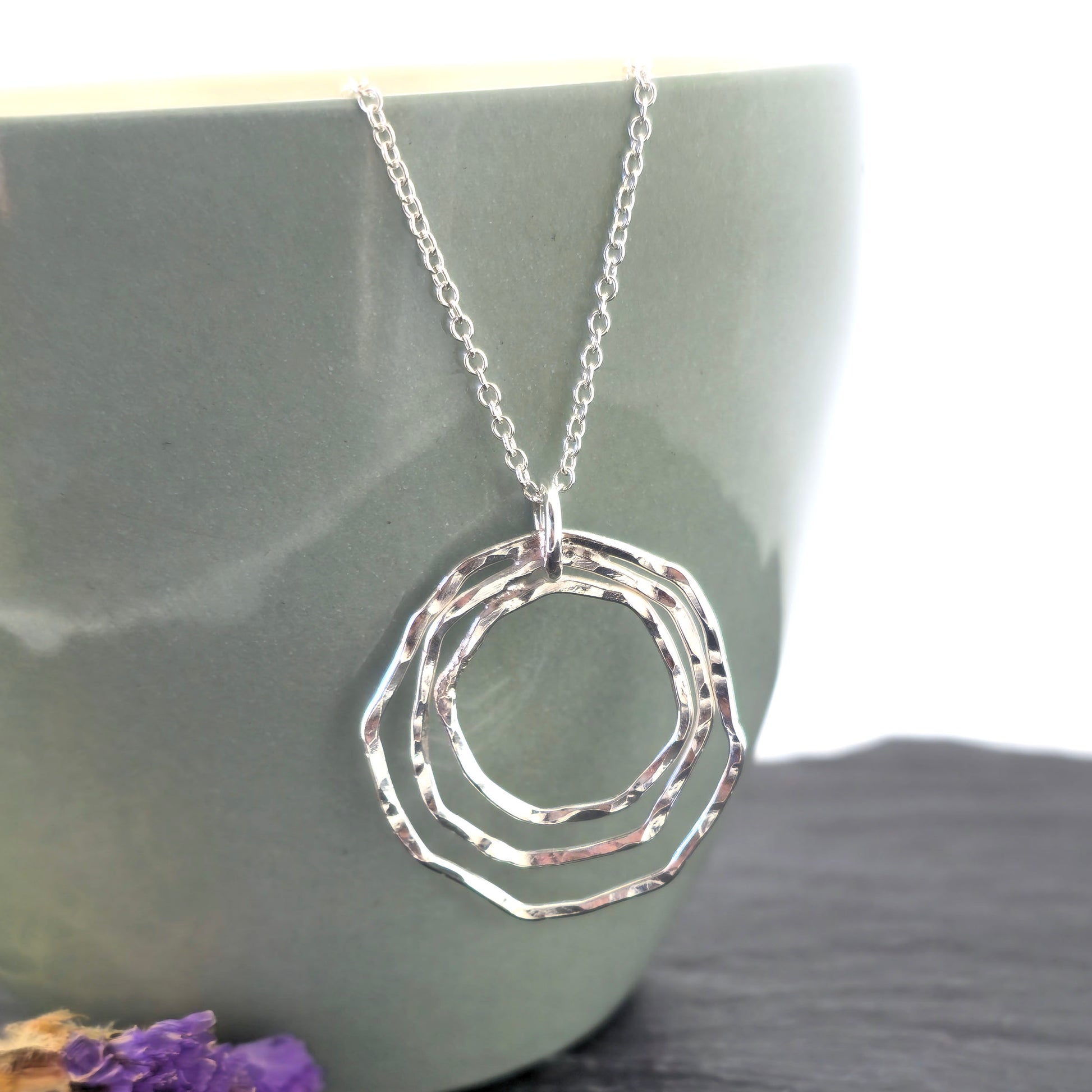 A handmade silver pendant consisting of three organic rustic style open circles of different sizes suspended from a silver chain. On a green cup with dried flowers. Mijoux Jewellery.