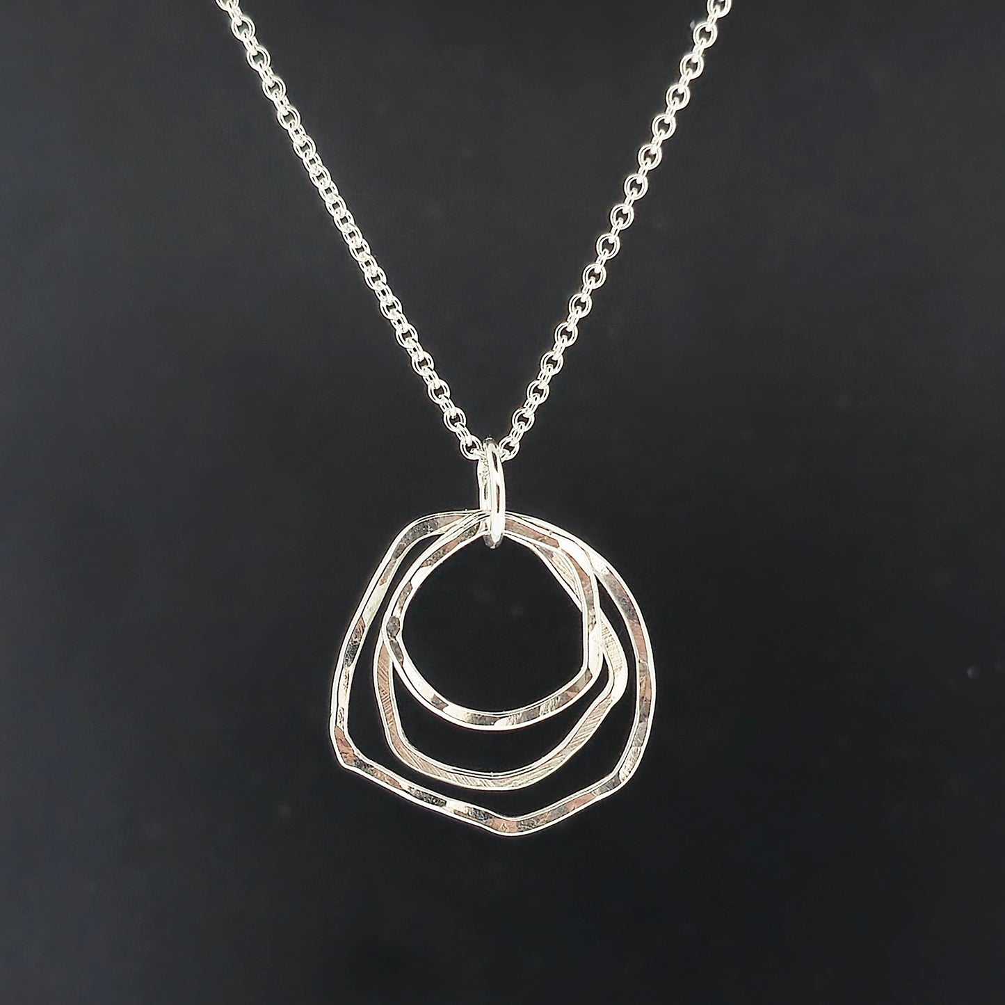 A silver pendant consisting of three organic rustic style circles of different sizes suspended together from a silver chain. On a black background.