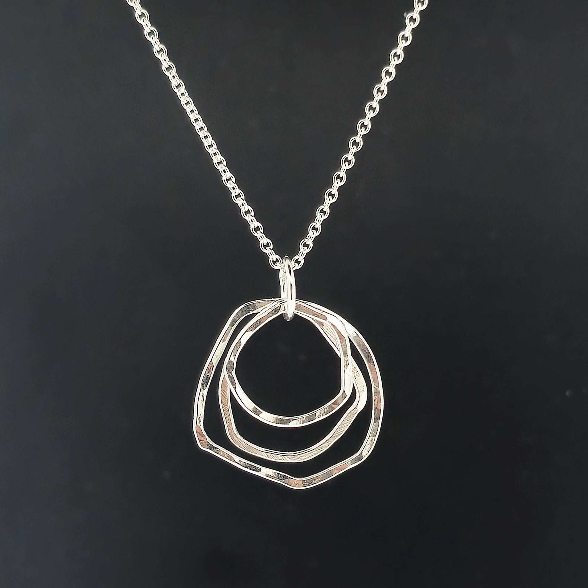 A silver pendant consisting of three organic rustic style circles of different sizes suspended together from a silver chain. On a black background.