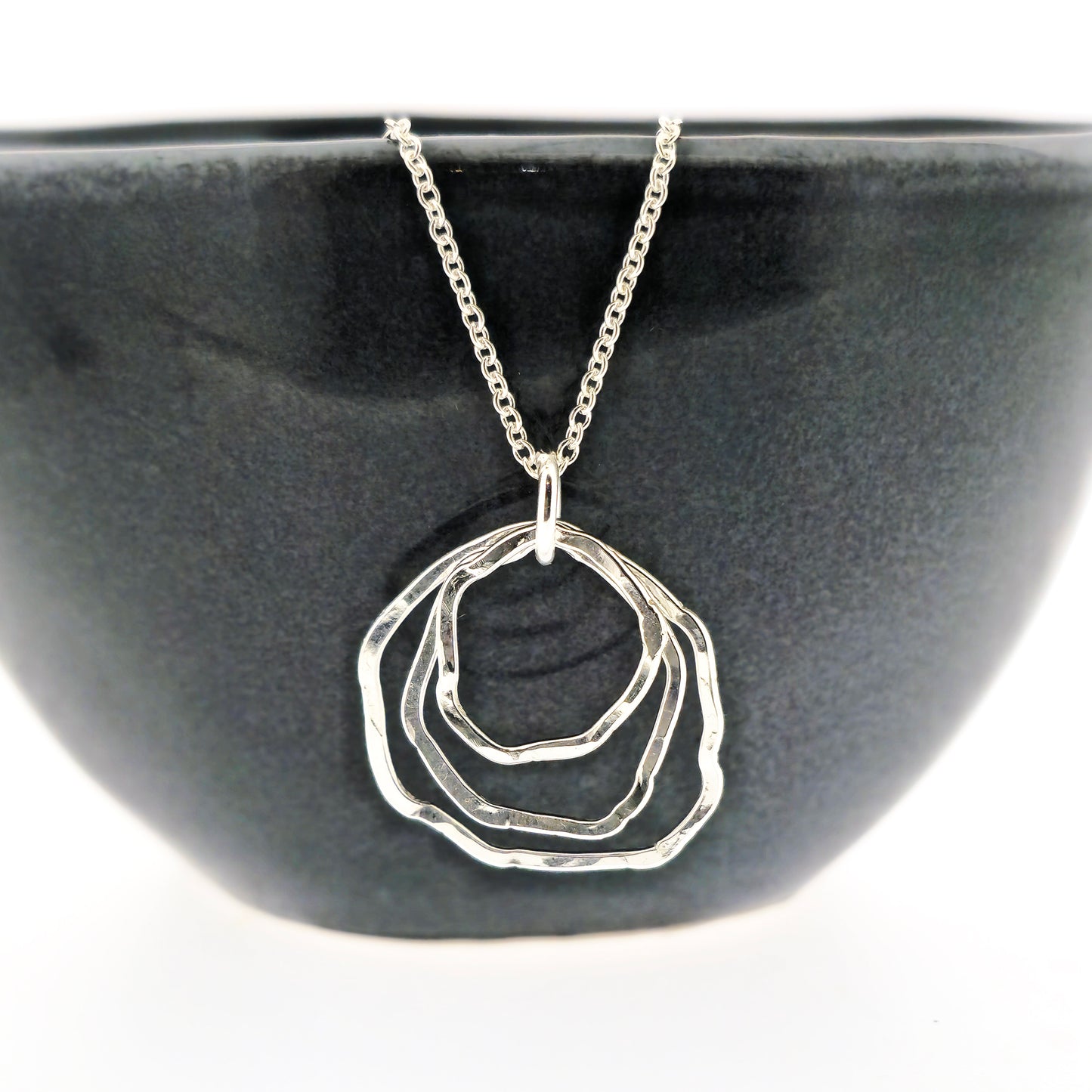 A silver pendant consisting of three organic rustic style circles of different sizes suspended together from a silver chain. Pictured on a black bowl.