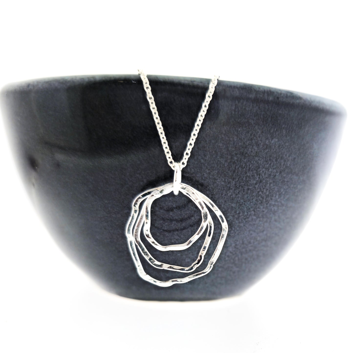 A silver pendant consisting of three organic rustic style circles of different sizes suspended together from a silver chain. Pictured on a black bowl.