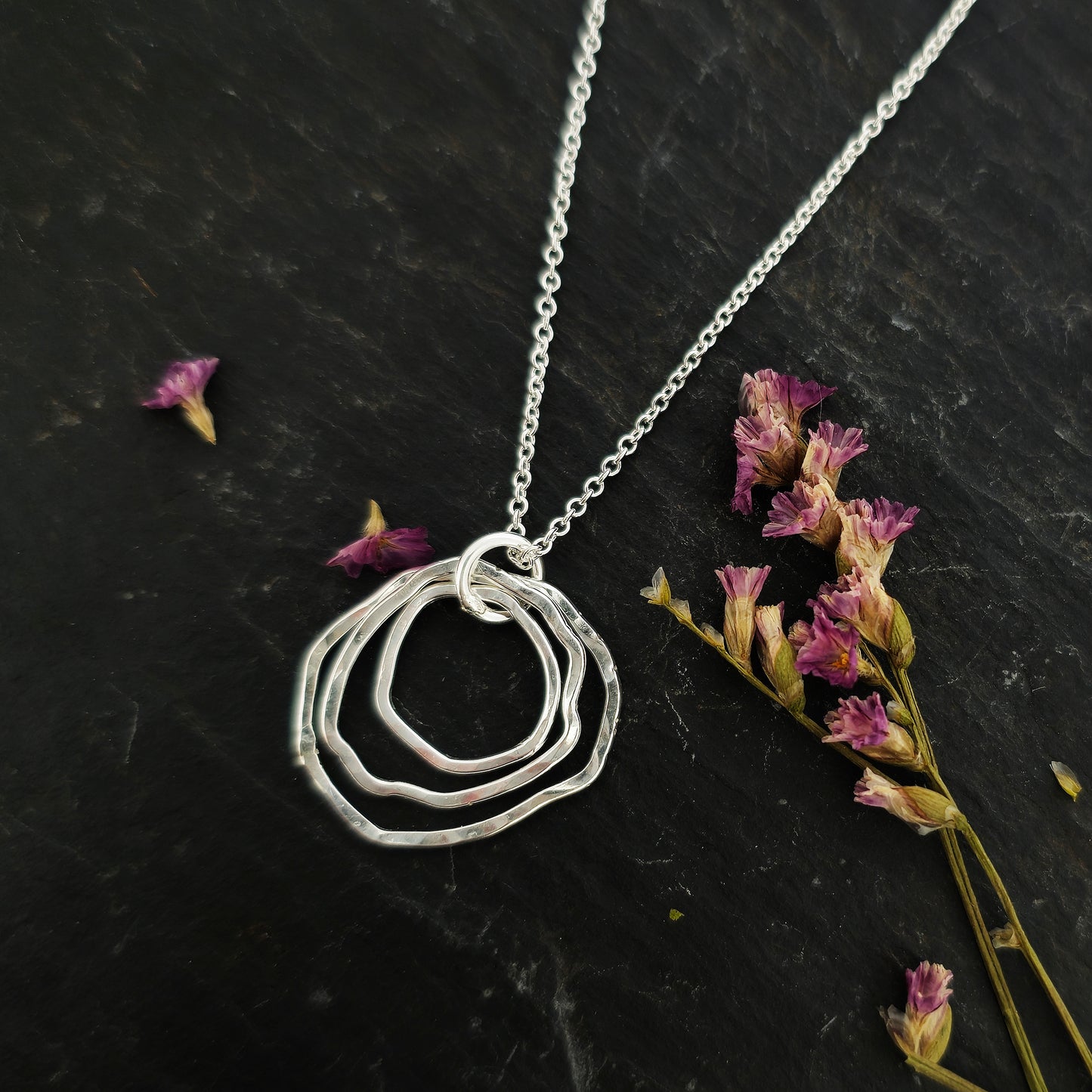 A silver pendant consisting of three organic rustic style circles of different sizes suspended together from a silver chain. Pictured on slate with flowers.