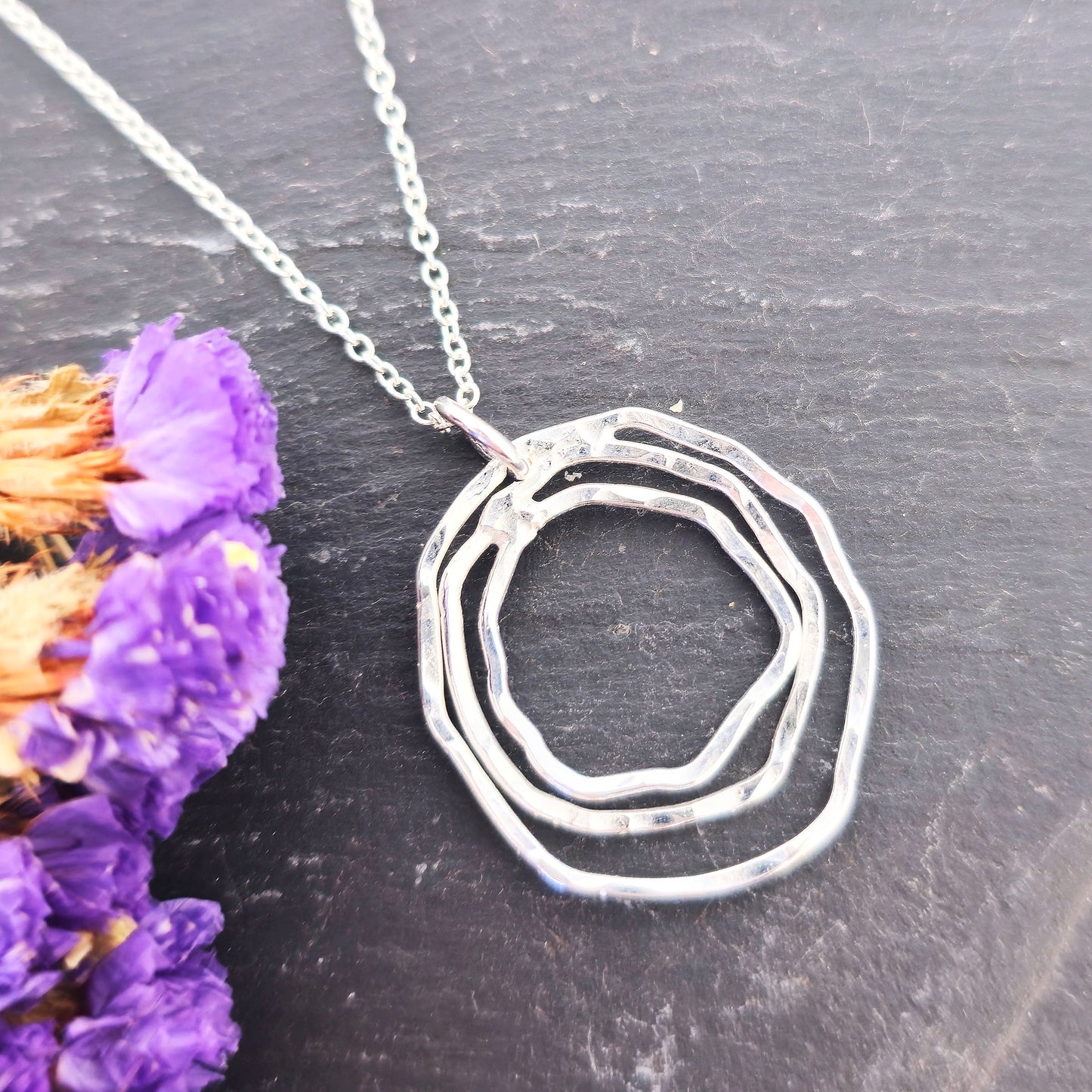 A handmade silver pendant consisting of two organic rustic style open circles of different sizes suspended from a silver chain. On a slate background with dried flowers. Mijoux Jewellery.