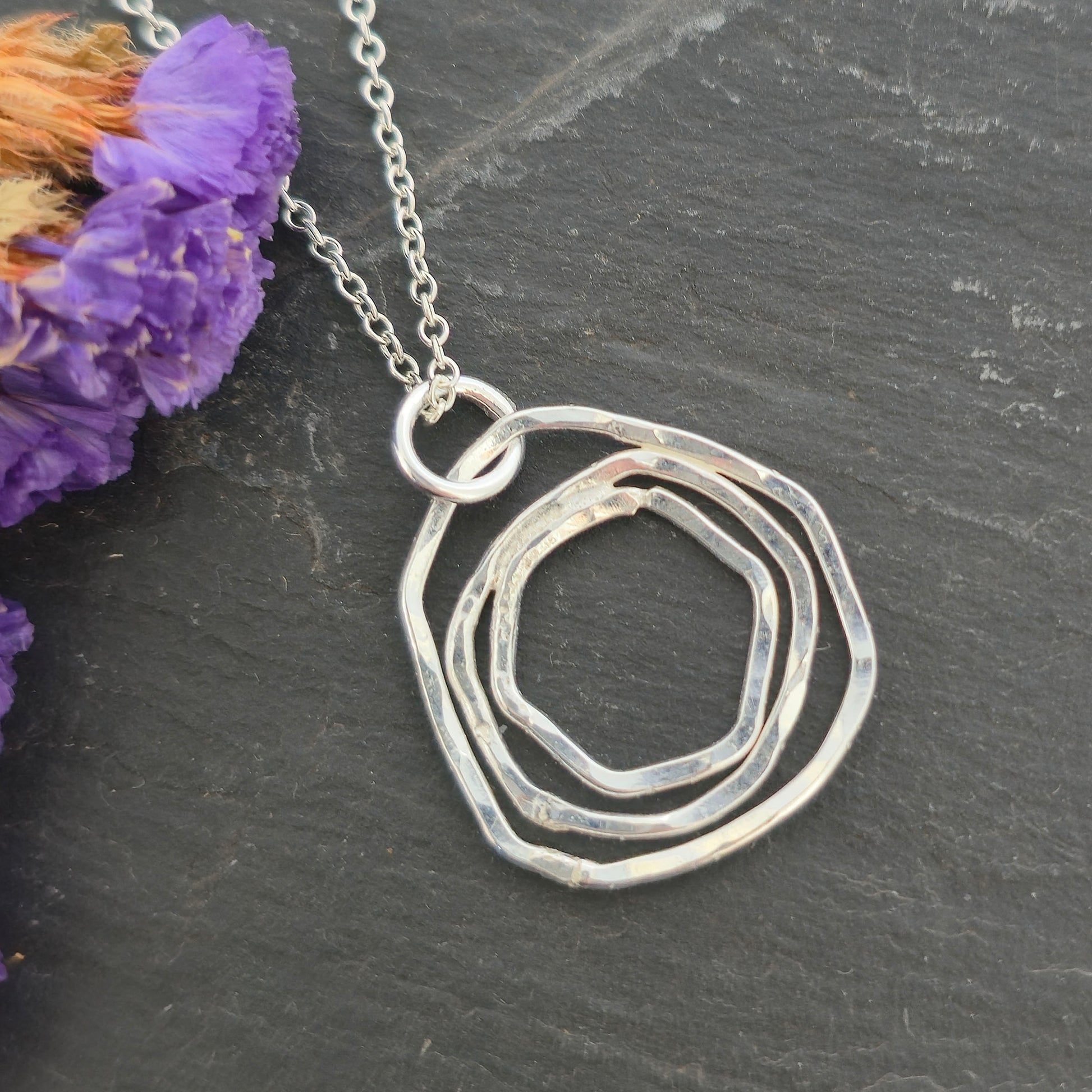 A silver pendant consisting of three organic rustic style circles of different sizes suspended from a silver chain. Pictured on a black background with purple flowers. Mijoux Jewellery.