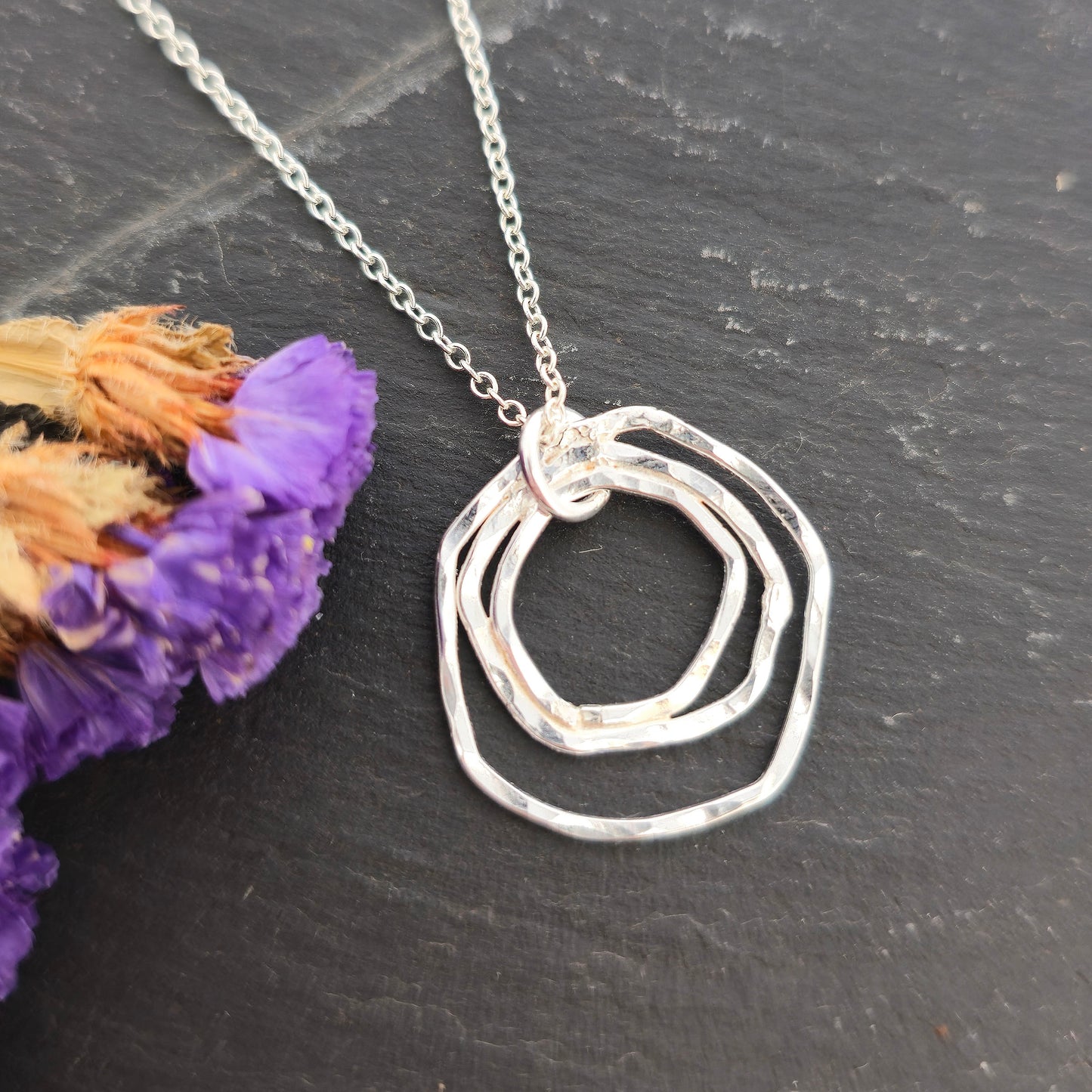 A silver pendant consisting of three organic rustic style circles of different sizes suspended from a silver chain. Pictured on a black background with purple flowers. Mijoux Jewellery.