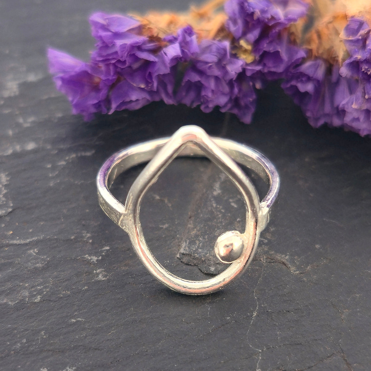 A handmade silver teardrop-shaped ring with a small ball detail, resting on a dark slate surface with purple dried flowers in the background. Mijoux Jewellery.