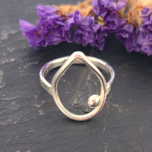 A handmade silver teardrop-shaped ring with a small ball detail, resting on a dark slate surface with purple dried flowers in the background. Mijoux Jewellery.