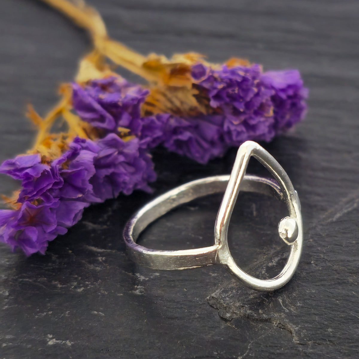 A handmade silver teardrop-shaped ring with a small ball detail, resting on a dark slate surface with purple dried flowers in the background. Mijoux Jewellery.