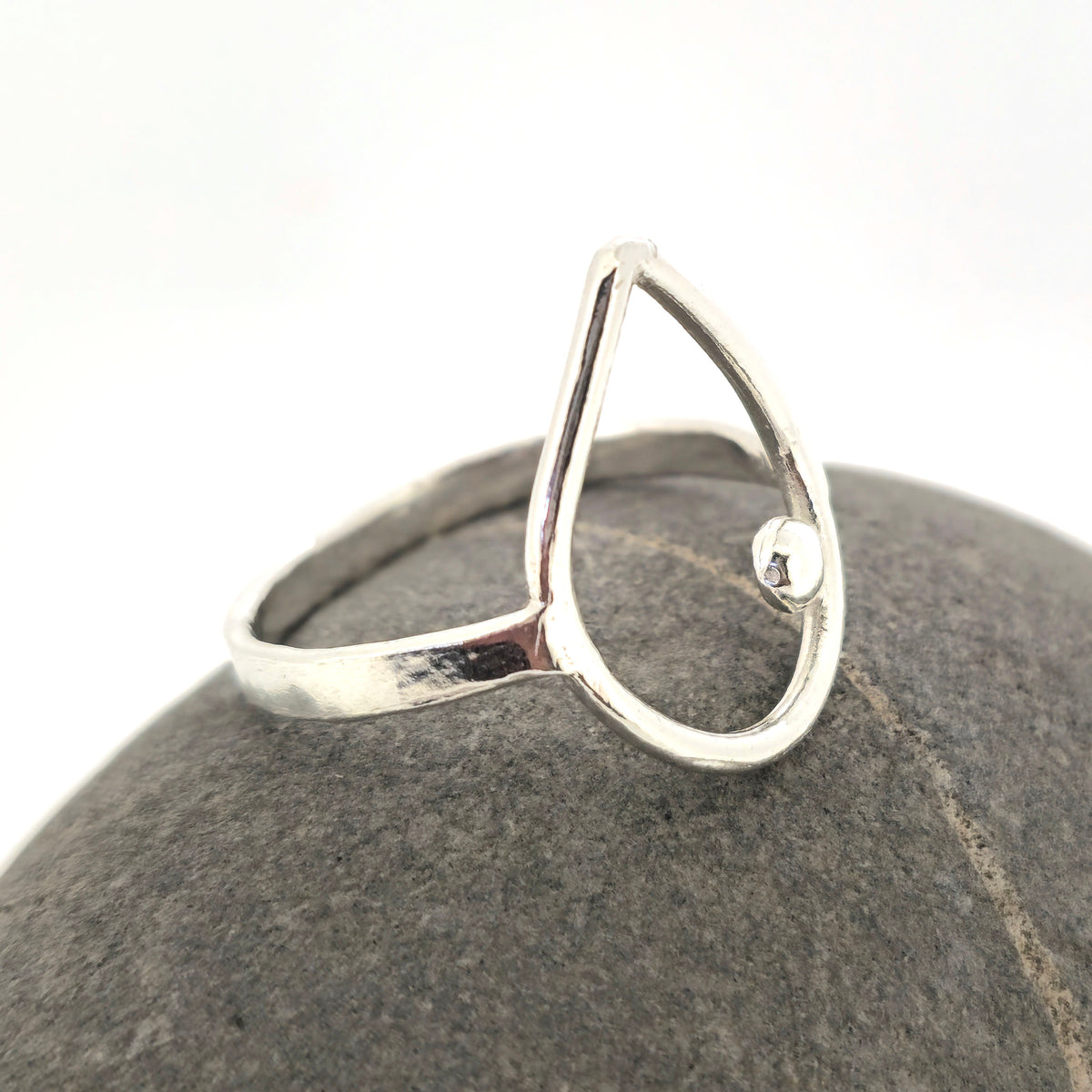 A handmade silver open teardrop ring with a small bell accent, displayed on a dark gray textured stone stand against a white background. Mijoux Jewellery.