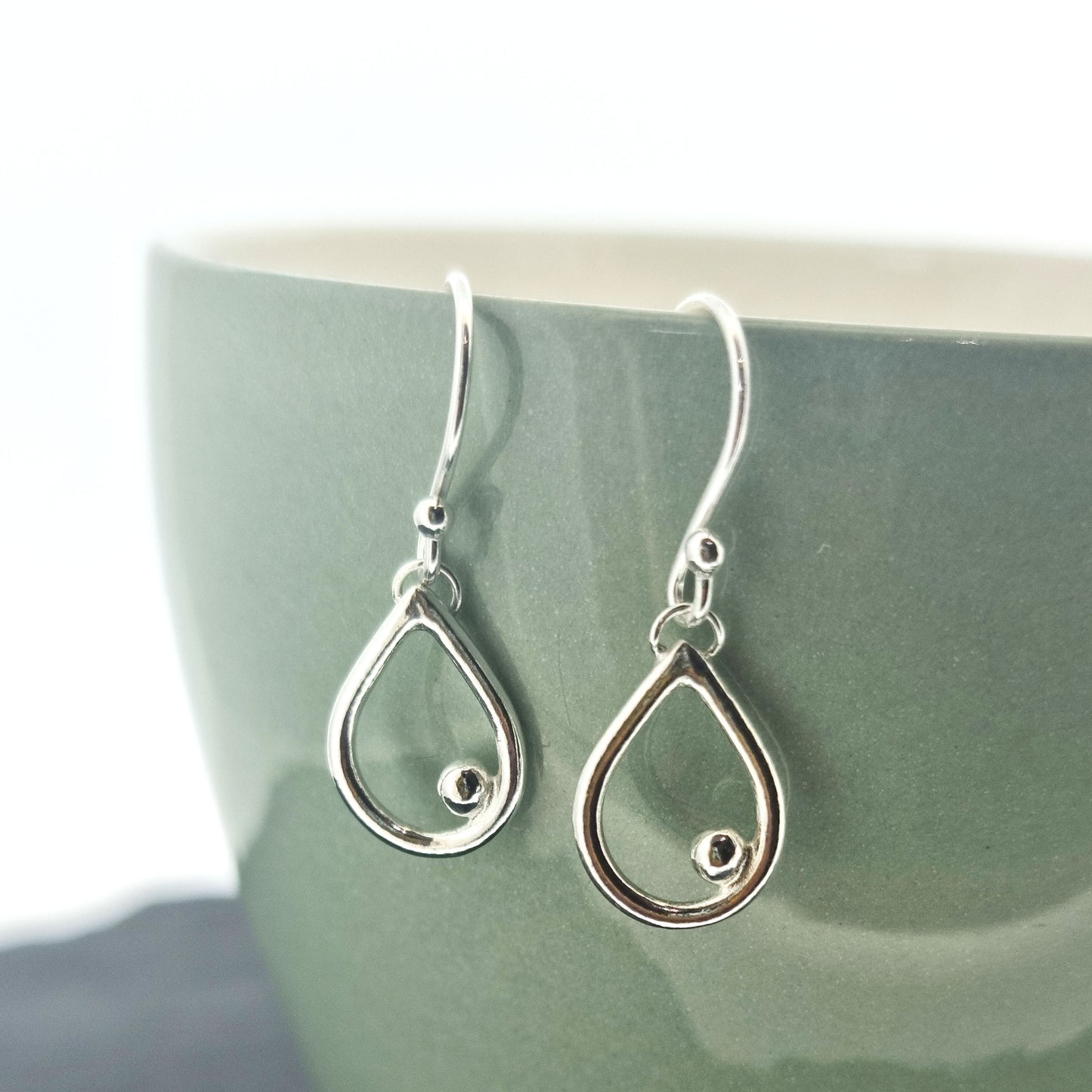 Silver drop earrings featuring an open teardrop shape with an off-centre ball. Small. Pictured on mug.