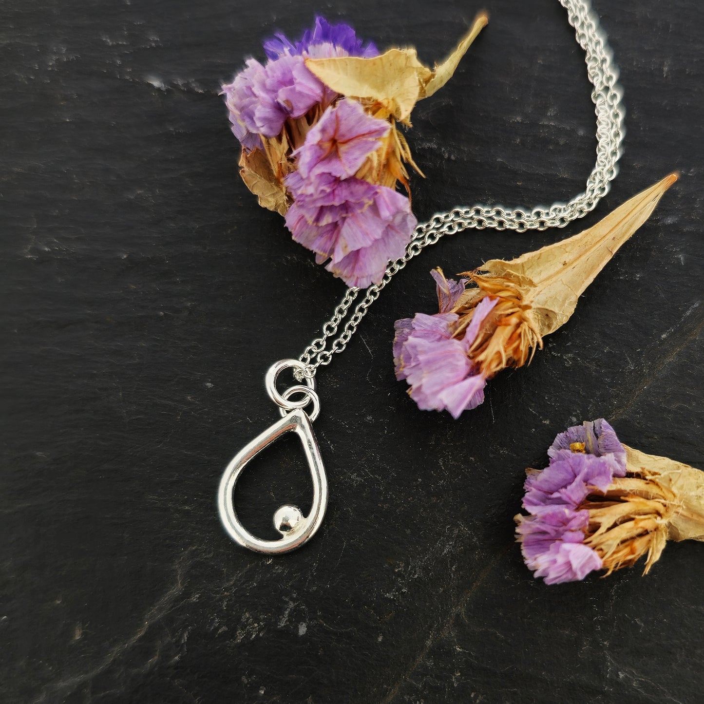 A silver open teardrop shaped pendant with an off-centre ball. On a silver chain. Small. Shown on slate with flowers.