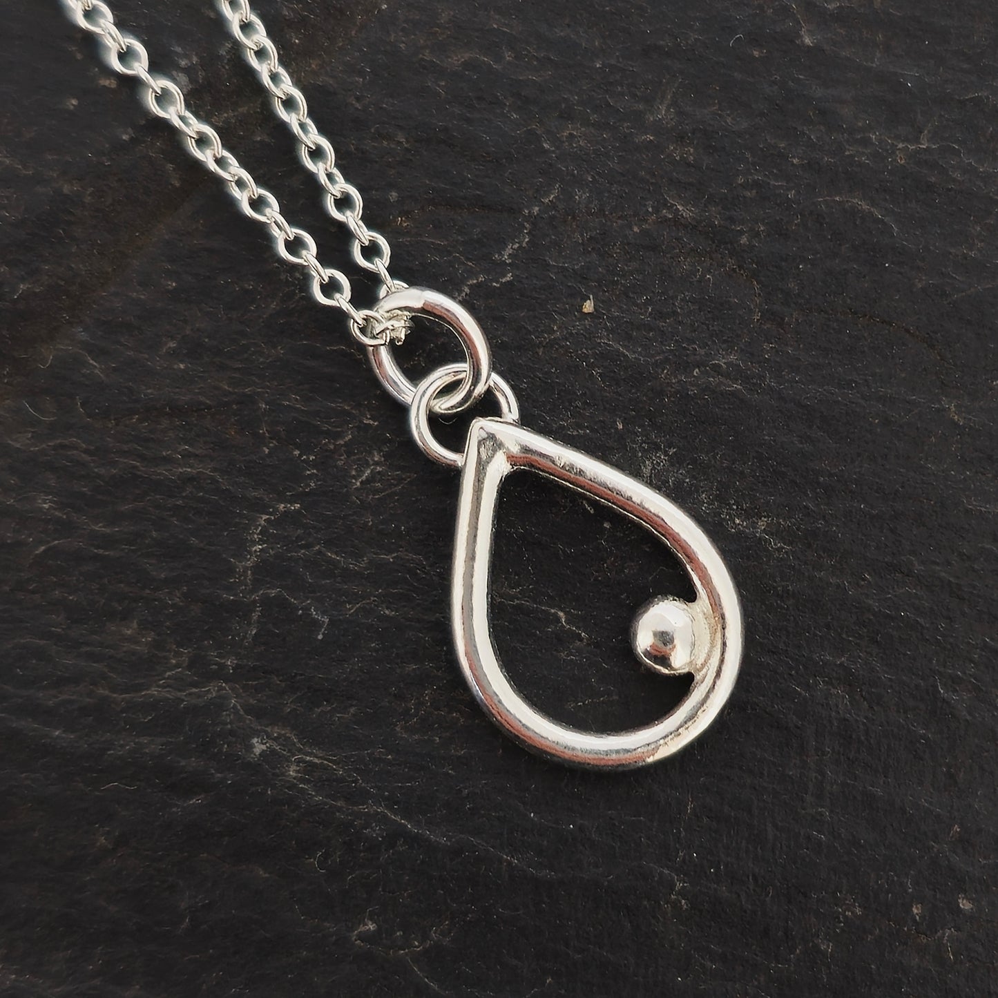 A silver open teardrop shaped pendant with an off-centre ball. On a silver chain. Small. Shown on slate.