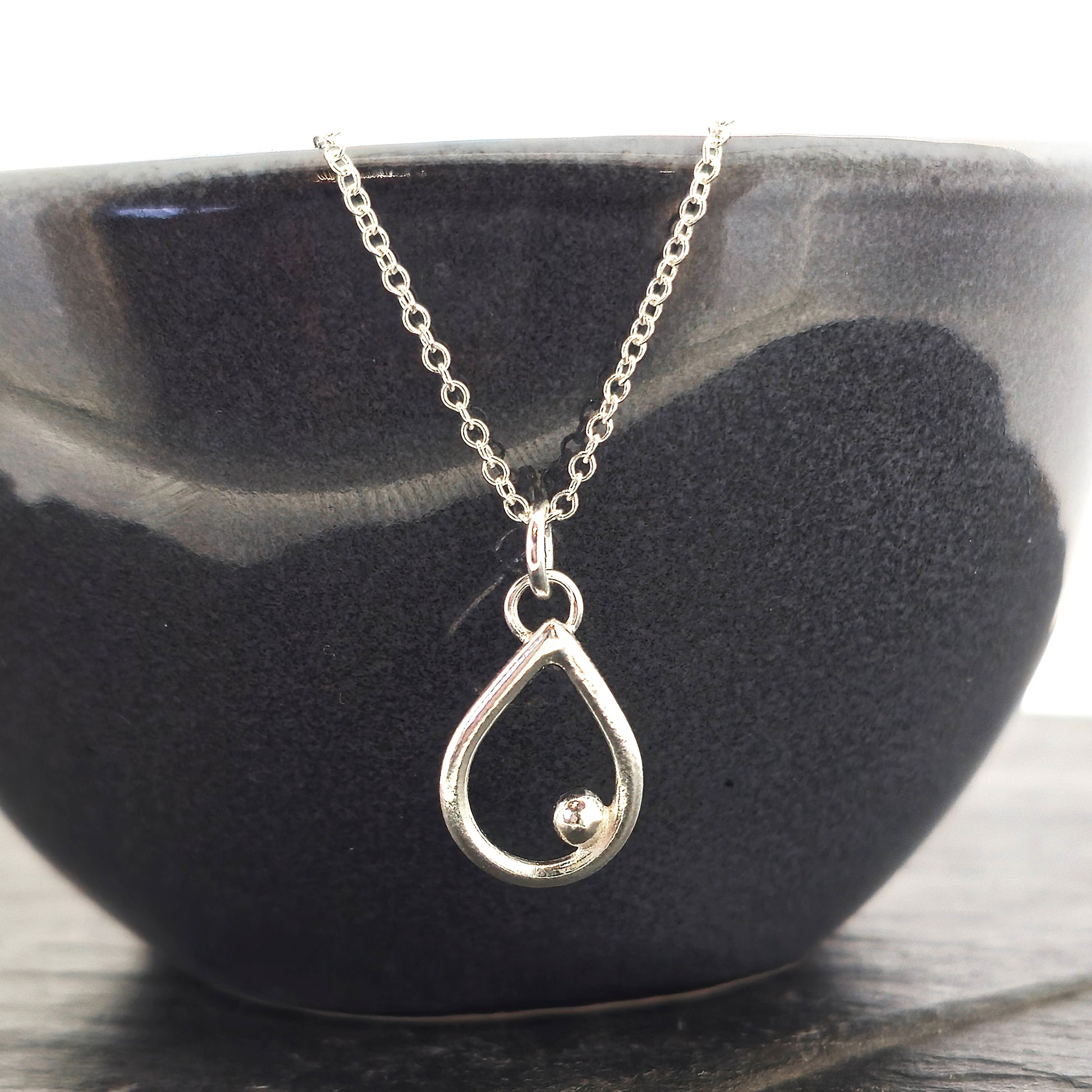 A silver open teardrop shaped pendant with an off-centre ball. On a silver chain. Small. Shown on a black bowl.