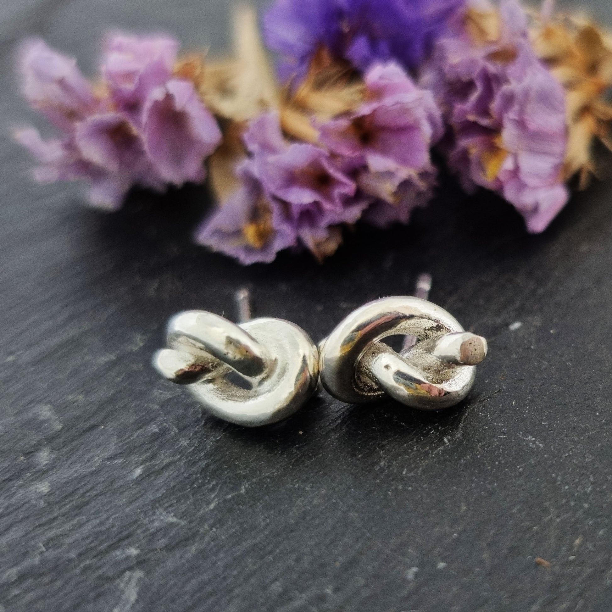 Silver stud earrings in the shape of a knot in a piece of string. Pictured with flowers.