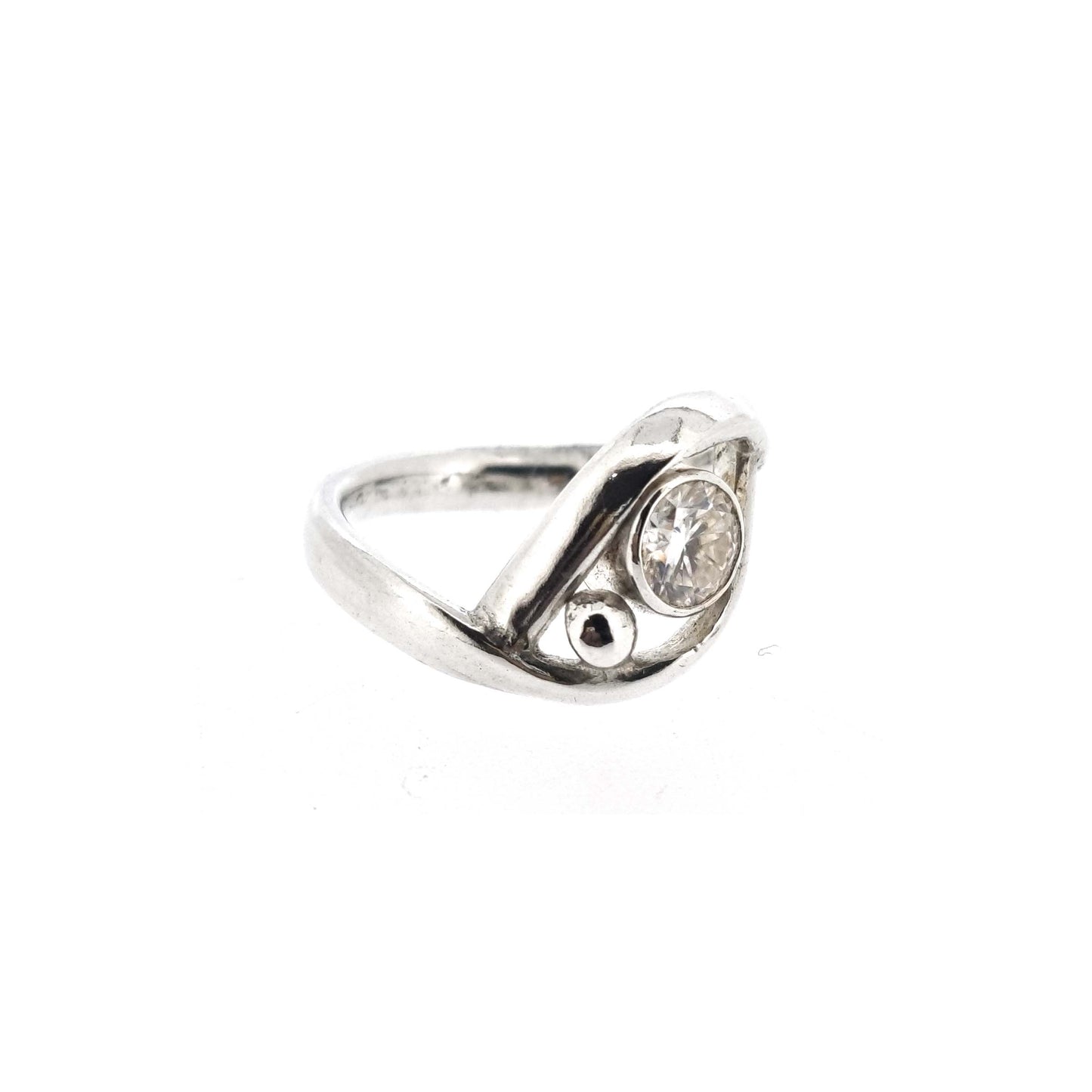 A silver ring with a central round faceted moissanite gemstone in a bezel setting. Next to the gemstone is a silver ball and the band of the ring wraps around both the ball and gemstone.