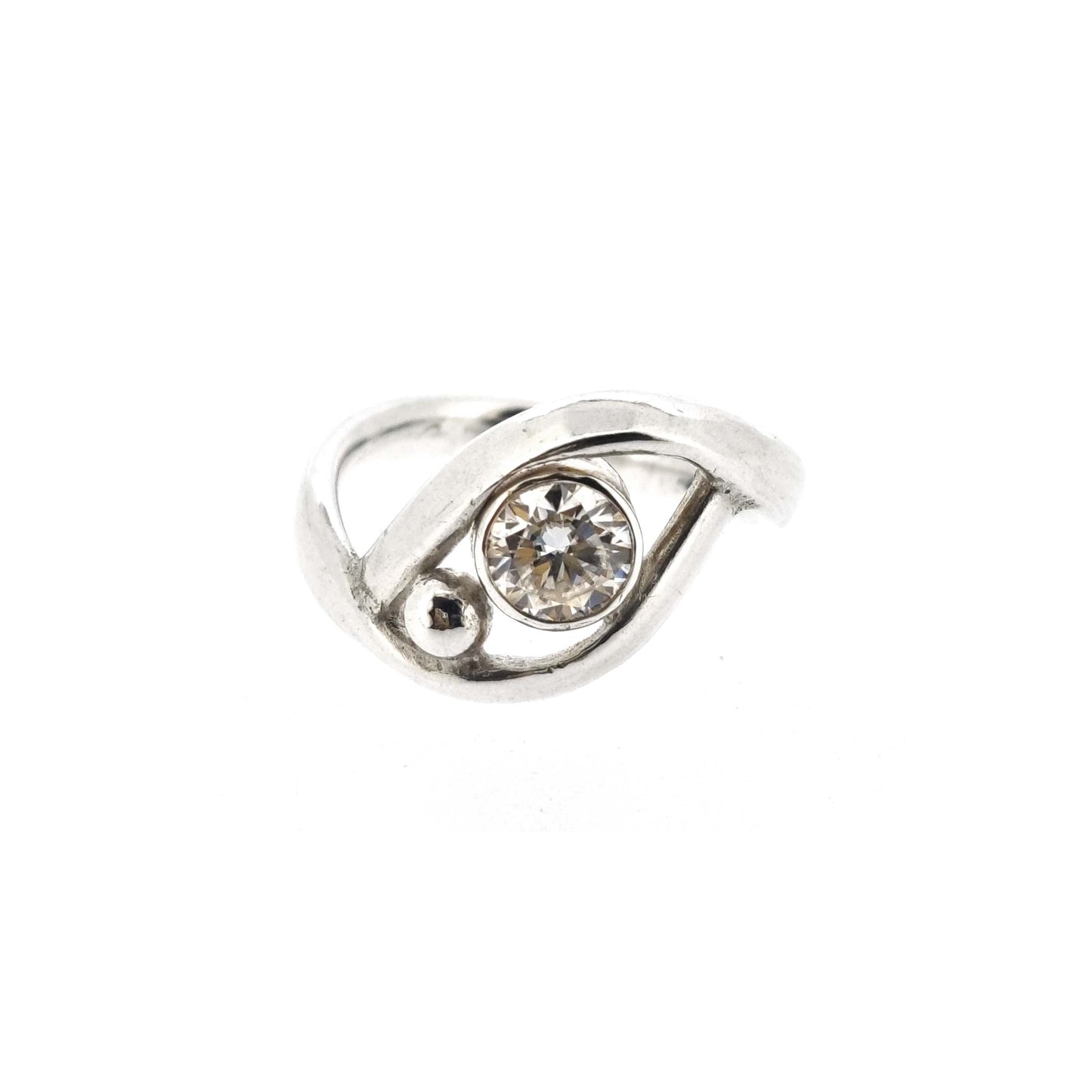 A silver ring with a central round faceted moissanite gemstone in a bezel setting. Next to the gemstone is a silver ball and the band of the ring wraps around both the ball and gemstone.