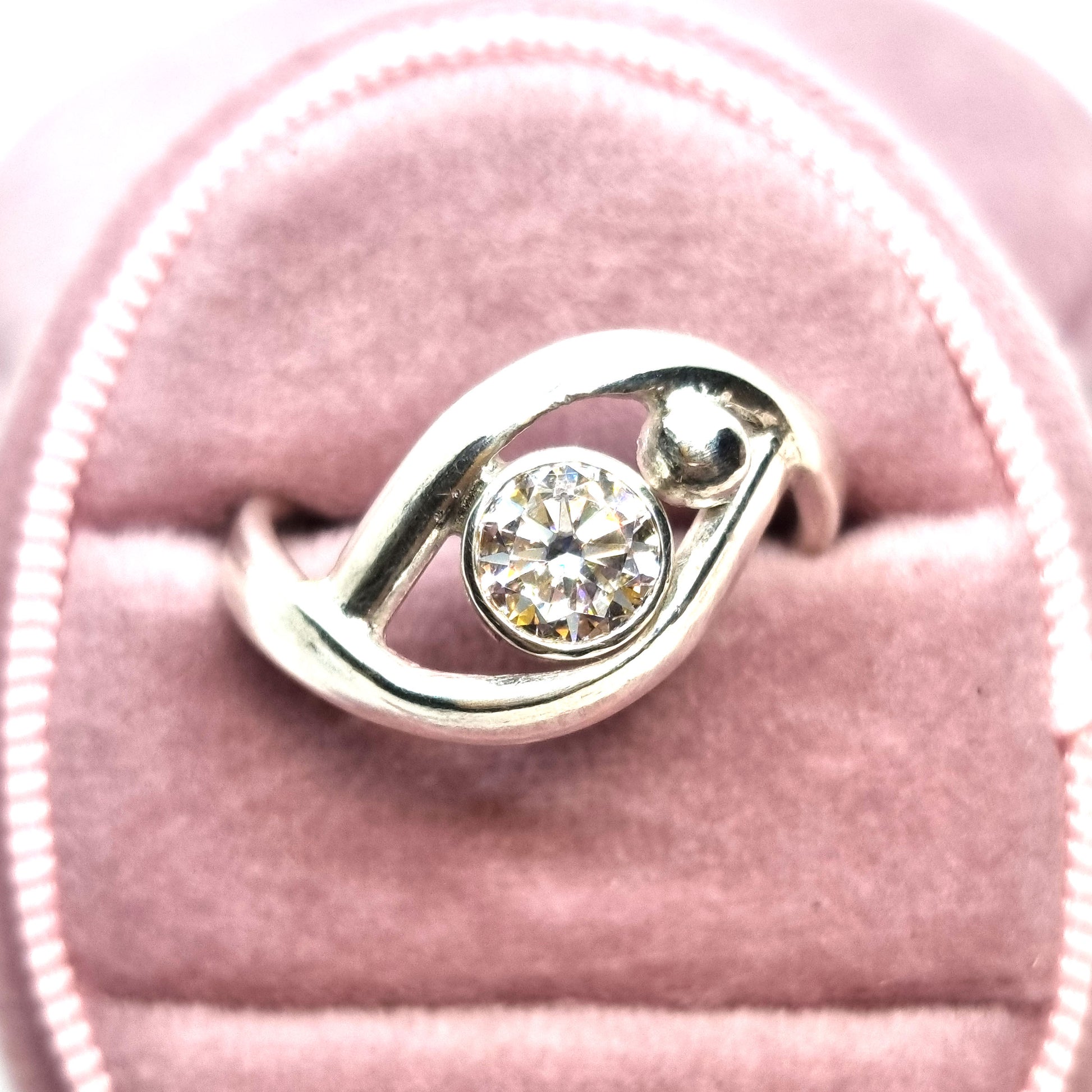 A silver ring with a central round faceted moissanite gemstone in a bezel setting. Next to the gemstone is a silver ball and the band of the ring wraps around both the ball and gemstone. Pictured in a pink jewellery box.