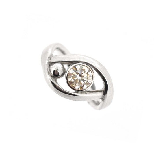 A silver ring with a central round faceted moissanite gemstone in a bezel setting. Next to the gemstone is a silver ball and the band of the ring wraps around both the ball and gemstone.