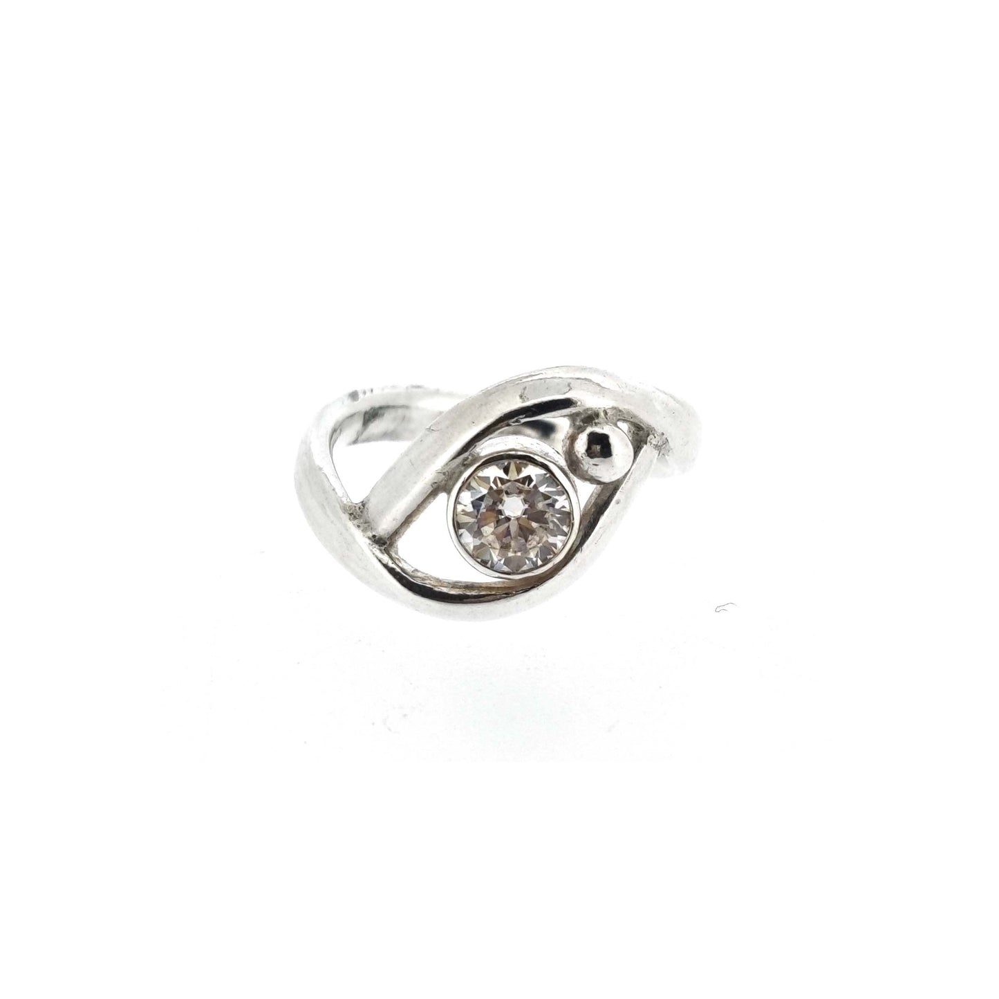 A silver ring with a central round faceted moissanite gemstone in a bezel setting. Next to the gemstone is a silver ball and the band of the ring wraps around both the ball and gemstone.