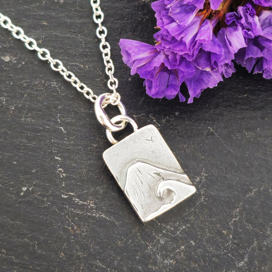 A rectangular silver handmade pendant featuring a mountain with a sea and wave in front and birds flying in the sky. Suspended from a silver chain. On a slate background with purple flowers. Mijoux jewellery.