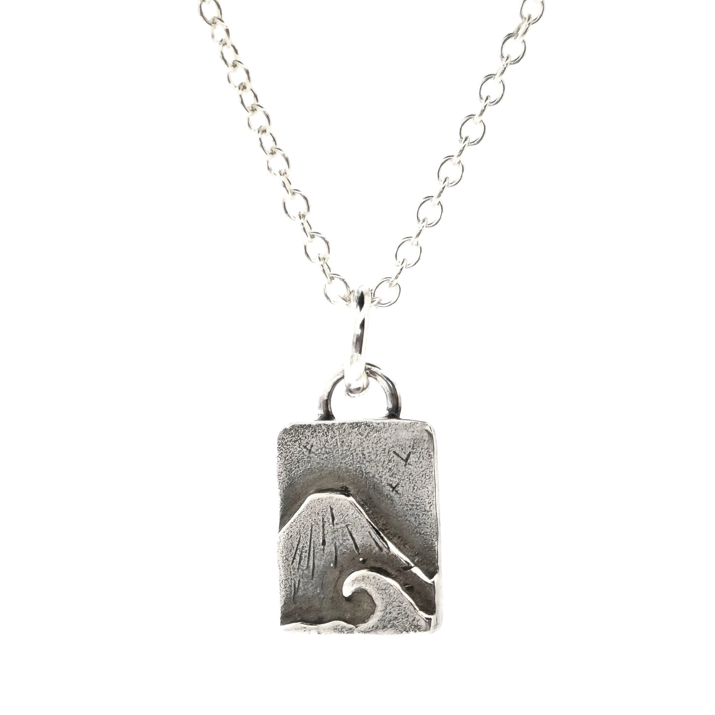 A rectangular silver handmade pendant featuring a mountain with a sea and wave in front and birds flying in the sky. Suspended from a silver chain. On a white background. Mijoux jewellery.