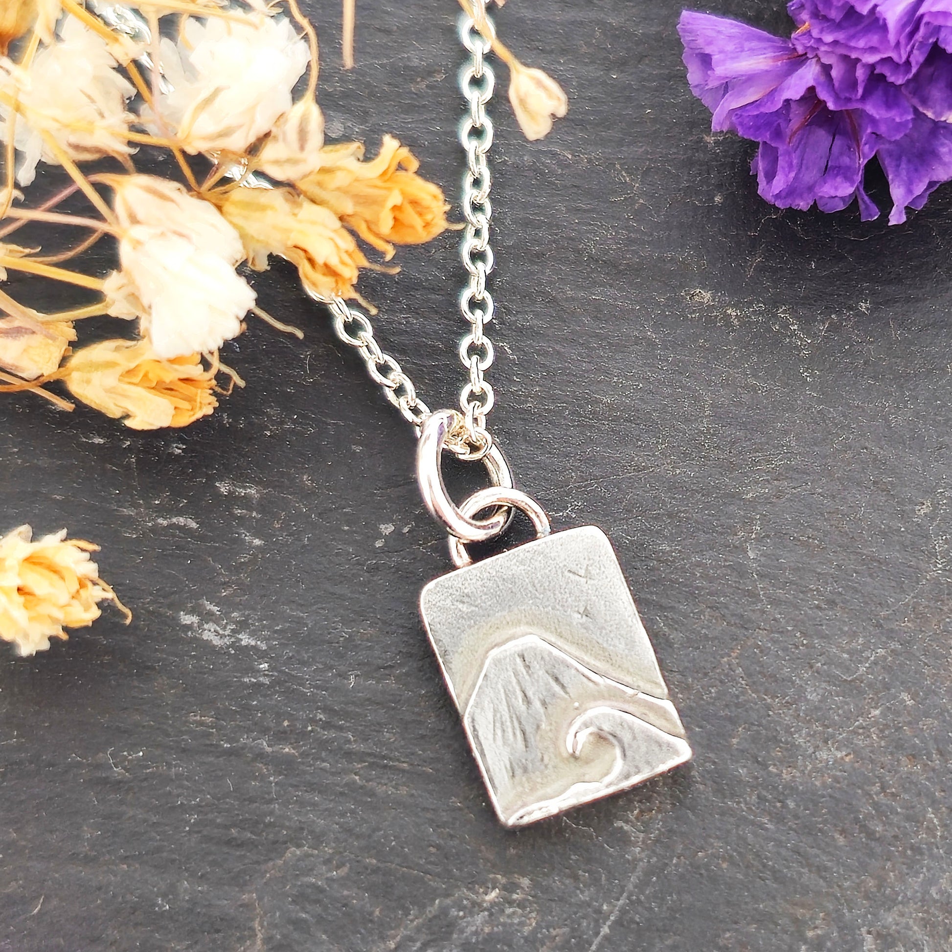 A rectangular silver handmade pendant featuring a mountain with a sea and wave in front and birds flying in the sky. Suspended from a silver chain. On a slate background with purple & white flowers. Mijoux jewellery.