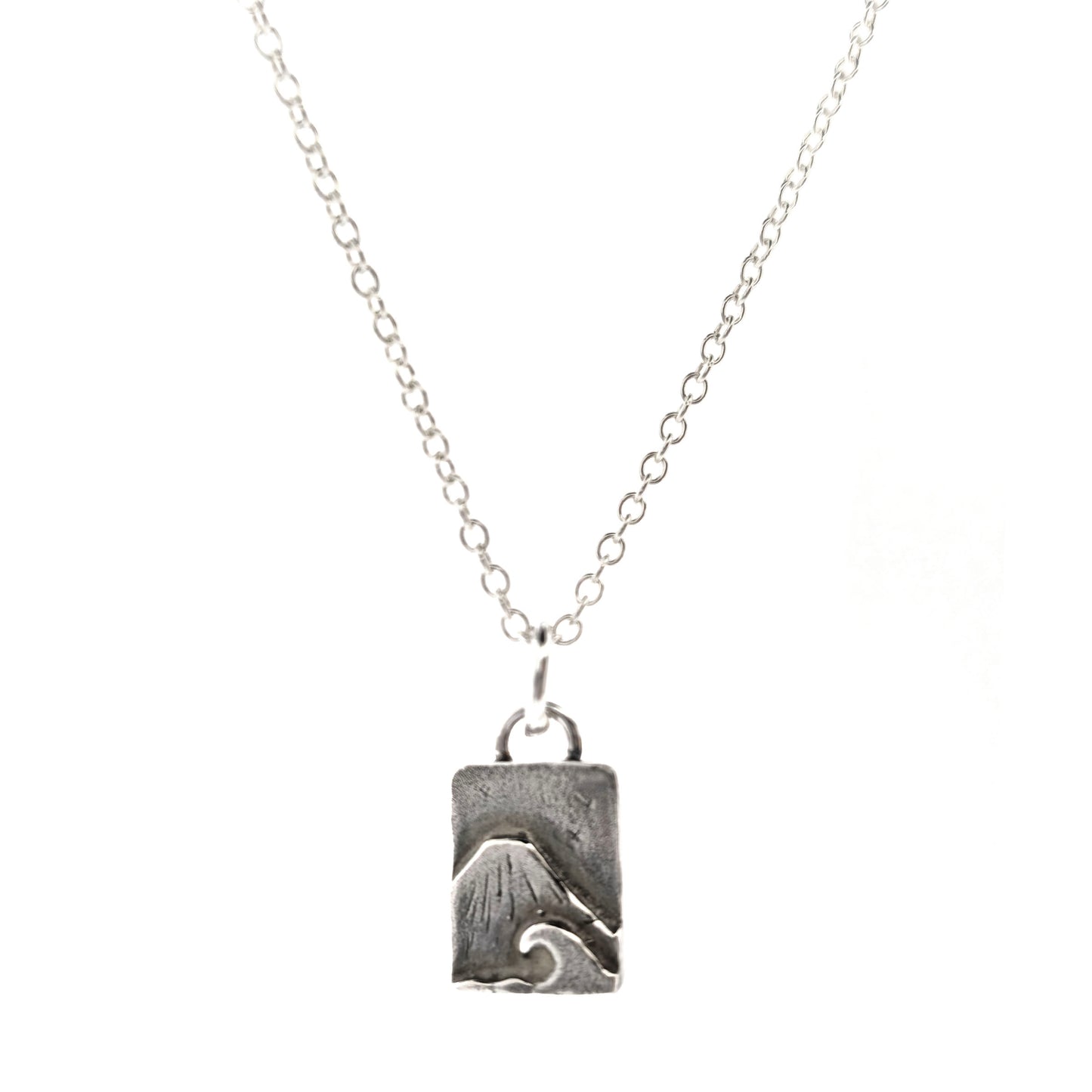 A rectangular silver handmade pendant featuring a mountain with a sea and wave in front and birds flying in the sky. Suspended from a silver chain. On a white background. Mijoux jewellery.