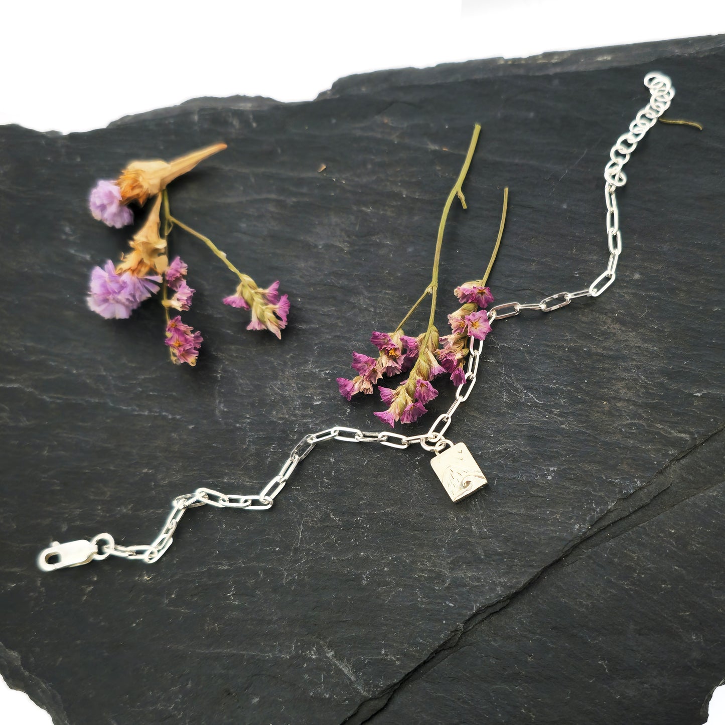 An silver bracelet with oblong links and a rectangular charm featuring a mountain and wave scene. Shown on slate with flowers.