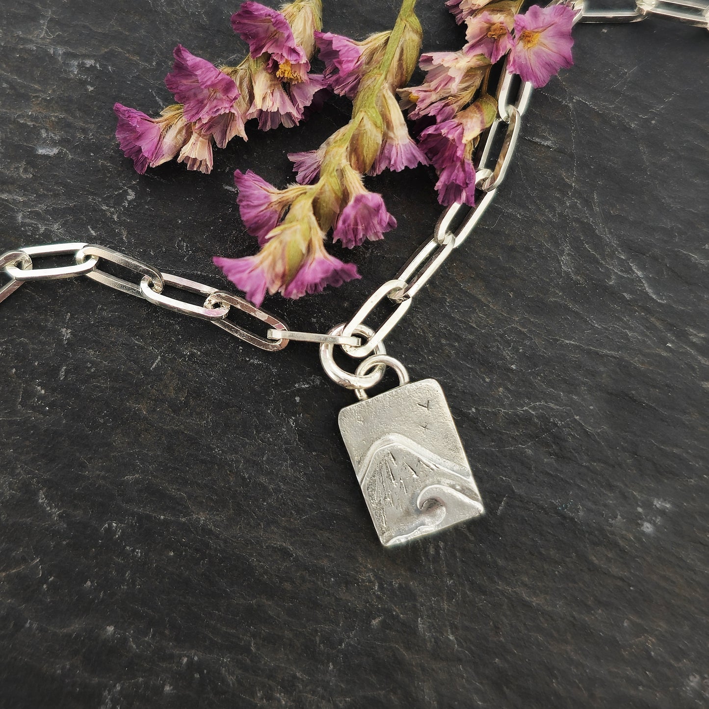 An silver bracelet with oblong links and a rectangular charm featuring a mountain and wave scene. Shown on slate with flowers.