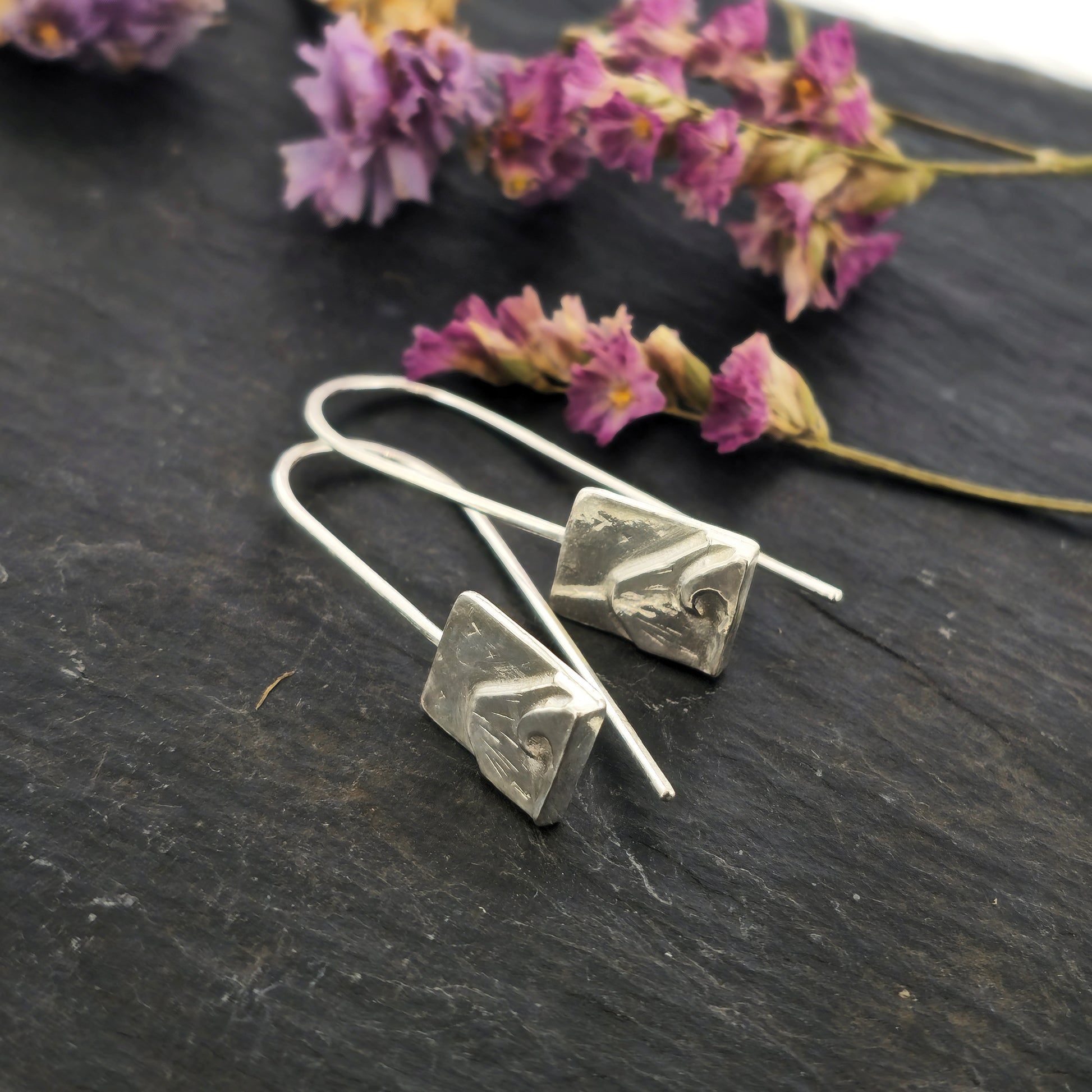 A pair of silver drop earrings featuring a rectangular section with a mountain and sea scene with birds on an ear wire. Shown on slate with flowers.