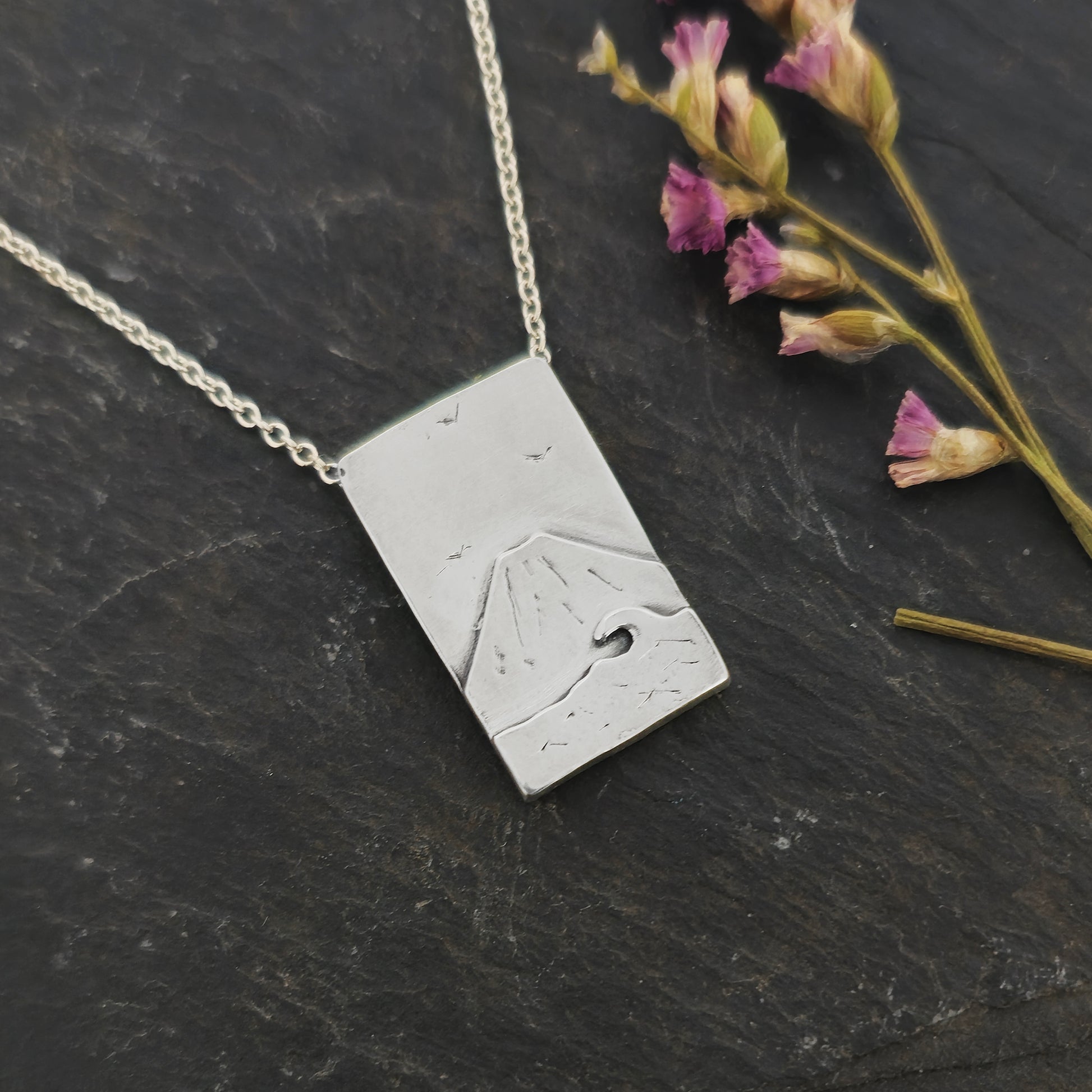 A rectangular silver pendant with a mountain scene featuring Mount Fuji behind a sea with a wave and birds in the sky. On a silver chain with invisible bail. Mijoux Creations. hown on slate with flowers.